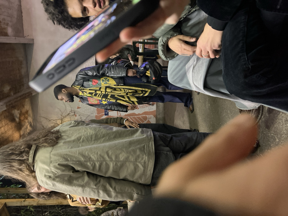 An indoor scene depicts a social gathering at a venue in Bagnolet, France, likely Alucinari, based on the visible "ALU" text on a mural-decorated wall and the industrial aesthetic. The perspective is from a low angle, looking up into the group.
In the immediate foreground, a blurred hand, holding a black smartphone, is visible on the lower right. To its left, a person is seated, their hands resting on their lap. They wear light grey sweatpants over dark trousers, and a silver ring is present on their left ring finger. Brown lace-up shoes are visible on their feet. Partially visible to the right of the phone is a person with dark, curly hair and light skin, looking towards the camera with a neutral expression.
In the mid-ground, a person with long, wavy brown hair stands with their back to the camera, wearing an olive-green jacket over dark pants. They face towards the left side of the frame. To their right, a Black male stands, facing left. He has a trimmed beard and short dark hair. He wears a black leather jacket adorned with multiple patches, including a yellow peace sign, a red heart, a yellow star, and a yellow and black shield design. A long, fringed black and yellow patterned scarf is draped around his neck. He wears dark blue flared trousers and brown shoes, holding a black object, possibly a small bag or helmet, in his left hand.
The background features light-colored walls with a faded, reddish-brown mural depicting abstract figures. Exposed wooden beams and a raw concrete ceiling are visible overhead. The floor is concrete. In the far left background, blurred green foliage is present, suggesting plants within the space or an adjacent outdoor area. Other individuals are vaguely visible in the background, some seated at tables, indicating a social event. The lighting is dim and artificial.