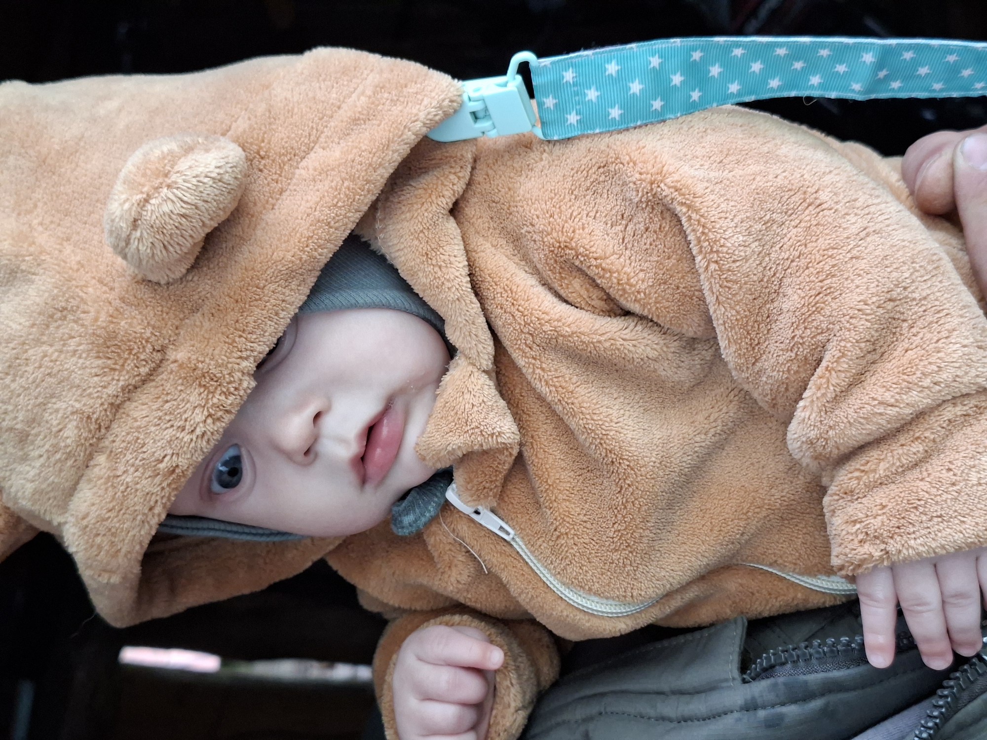 A close-up image of a baby wearing a brown, fuzzy bear-eared hooded jacket and a gray knit cap, lying in a stroller in Unknown, Unknown. The baby has blue eyes and a slightly open mouth. A blue strap with white stars is visible. The baby's hands are visible against a gray-green surface.