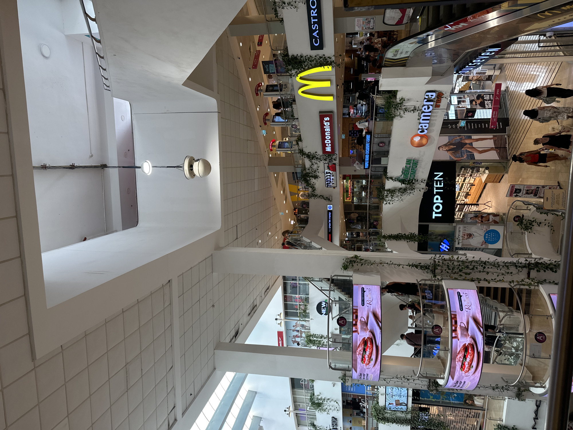 This is a vertical, eye-level shot of the interior of a modern shopping mall in Tel Aviv, Israel. The scene is filled with a variety of shops and advertisements, suggesting a busy commercial environment.
Dominating the upper portion of the image is a large, bright yellow McDonald's "M" logo, with the "McDonald's" name visible in red below it. To the left of the McDonald's sign, a "CASTRO" sign is visible, also illuminated. Further down, a store called "camera" is advertised with its logo, and below that, a store named "TOPTEN" is prominently displayed. Several other shop names and advertisements are visible, including "PURPLE BEARD EXCLUSIVE T-SHIRT" and "AMERICAN COMFORTY".
Escalators are visible, indicating different levels of the mall, and people can be seen walking around or using the escalators. Some individuals are casually dressed, suggesting a typical day of shopping. Green vines are draped along some of the store fronts and structural elements, adding a touch of greenery to the otherwise urban setting. Large digital screens display advertisements, one featuring images of food items and the "Romeo" brand.
The mall appears to be well-lit, with a combination of artificial lighting and natural light filtering in from above through large windows or skylights. The overall impression is one of a vibrant and active shopping destination, likely during daytime hours. The visible text is predominantly in English and Hebrew, consistent with the location.