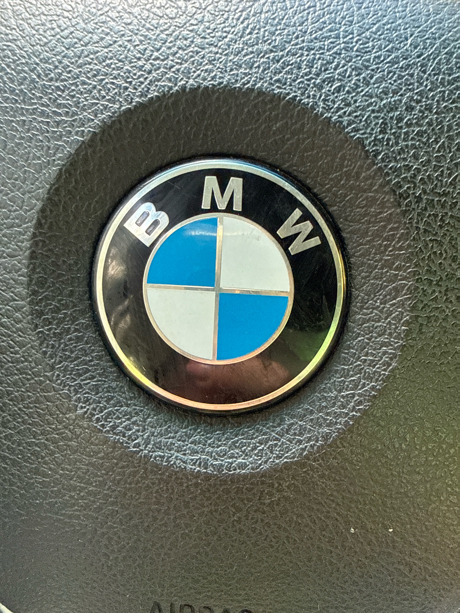 The image shows a close-up of a BMW logo on a steering wheel. The steering wheel is black and appears to be made of leather, with a textured surface. The BMW logo is round with a silver rim and a black background. Inside the logo, there is a circular emblem divided into four quadrants. Two opposite quadrants are white, and the other two opposite quadrants are blue. The letters "B," "M," and "W" are visible in silver on the black background of the logo, encircling the emblem. There is a shadow cast on the steering wheel to the left of the logo, indicating a light source from the right. The image is taken from a slightly elevated angle, looking down at the steering wheel. The overall impression is of a detail shot of a car's interior. No specific location within the city of Свенскі сельскі Савет, Belarus, can be determined from this image.