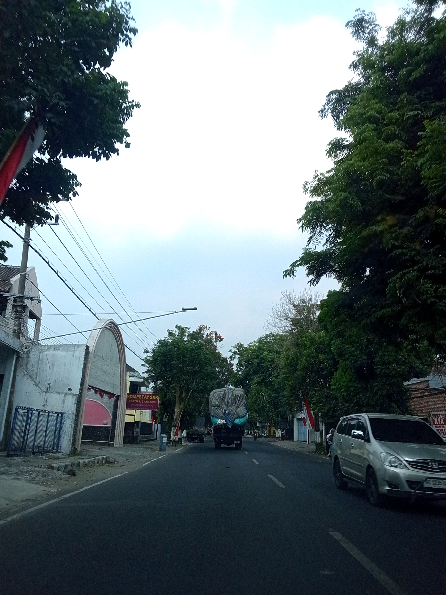 The image depicts a street scene in the city of Ngasem, Indonesia. The perspective is from a vehicle traveling on the road. On the left side of the road, there is a building with a white facade and a metal gate. A sign on the building reads "HOMESTAY SRK RESTO & CAFE SRK". Further down the road, there are trees and power lines overhead. A silver SUV is visible on the right side of the road, and a truck covered with a tarp is in the distance on the left side of the road. The sky is overcast.