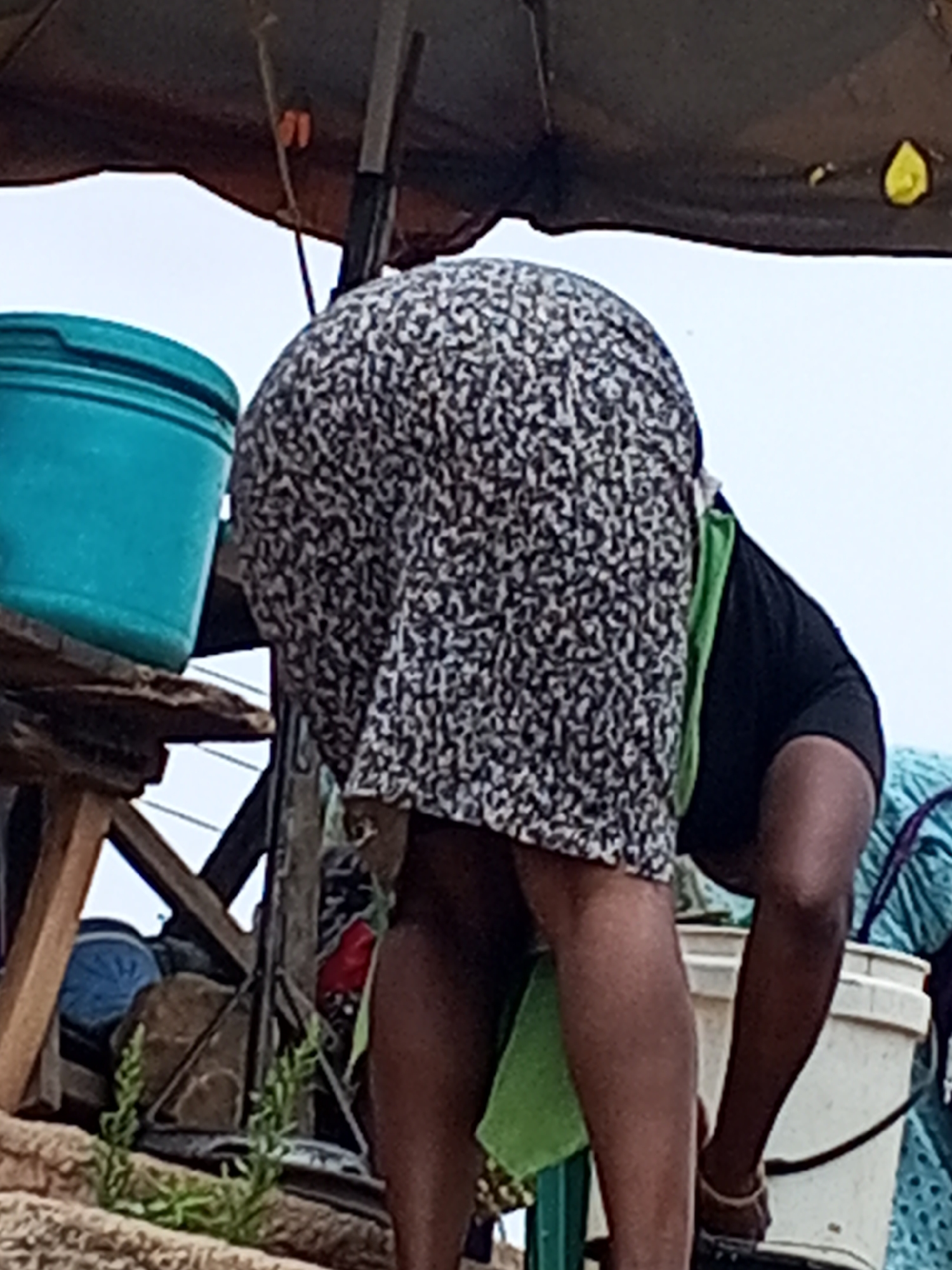 The image shows a woman bending over near a blue bucket in Gwong, Nigeria. She is wearing a black and white patterned dress, a black shirt, and sandals. A green cloth is draped over her shoulder. She is next to a white bucket. The environment includes a wooden structure and a cloth-covered frame. The location is implied to be Gwong, Nigeria. The main action is the woman bending over.