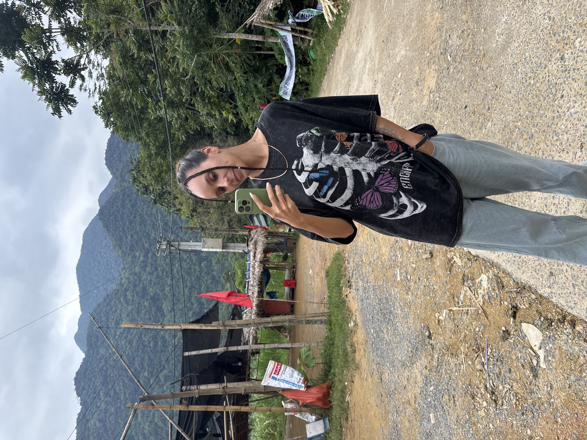 A woman stands on a gravelly dirt road in Cổ Lũng Commune, Vietnam, taking a selfie with her phone. She has dark braided hair and is wearing a black t-shirt with a skeleton graphic and light blue jeans. Her left hand is in her pocket, and her right hand holds the phone. In the background, there are thatched-roof huts, bamboo structures, lush green mountains, and trees under a cloudy sky. A red flag is visible on one of the huts.