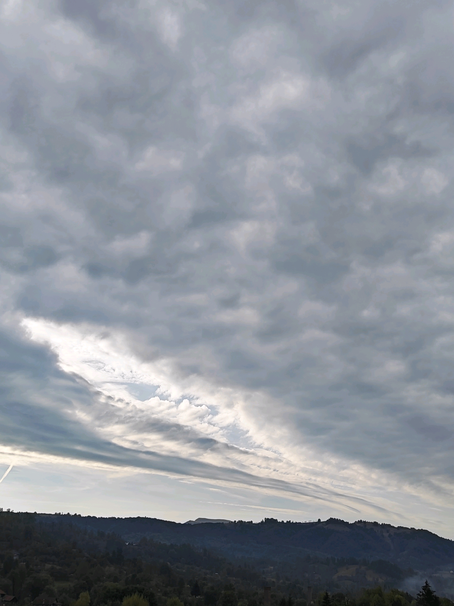 The image shows a landscape with a prominent mountain range in the foreground, covered in dark, dense vegetation. The sky above is filled with a complex pattern of gray clouds, with a bright, elongated opening revealing a lighter sky beneath. The overall impression is of a cloudy or overcast day. The scene appears to be a natural landscape, with no specific man-made structures or people clearly visible. The location is identified as Petroșani, Romania.