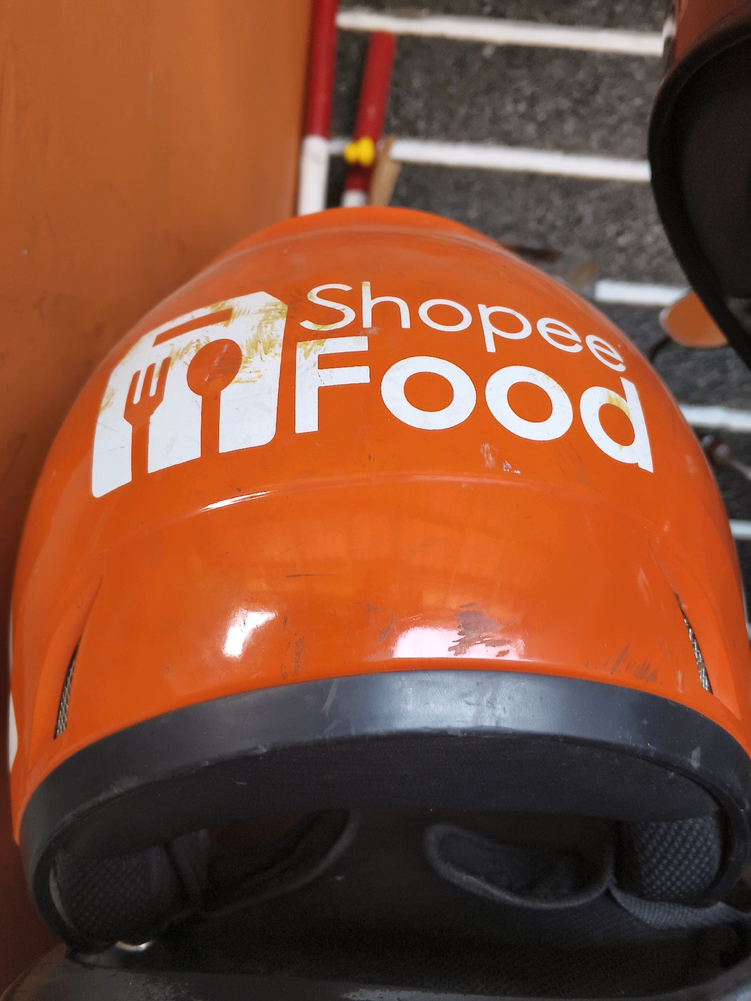 An orange Shopee Food helmet is shown up close, angled slightly to the left. The helmet has a white logo featuring a fork and spoon symbol next to the text "Shopee Food". The helmet appears to be used, with some scuff marks and scratches visible on its surface. The background is out of focus, showing a reddish-brown wall and a gray and white striped surface, possibly stairs or a walkway. Some red and yellow poles are also visible in the background, suggesting an outdoor setting. The image captures the details of the helmet, highlighting its branding and condition.