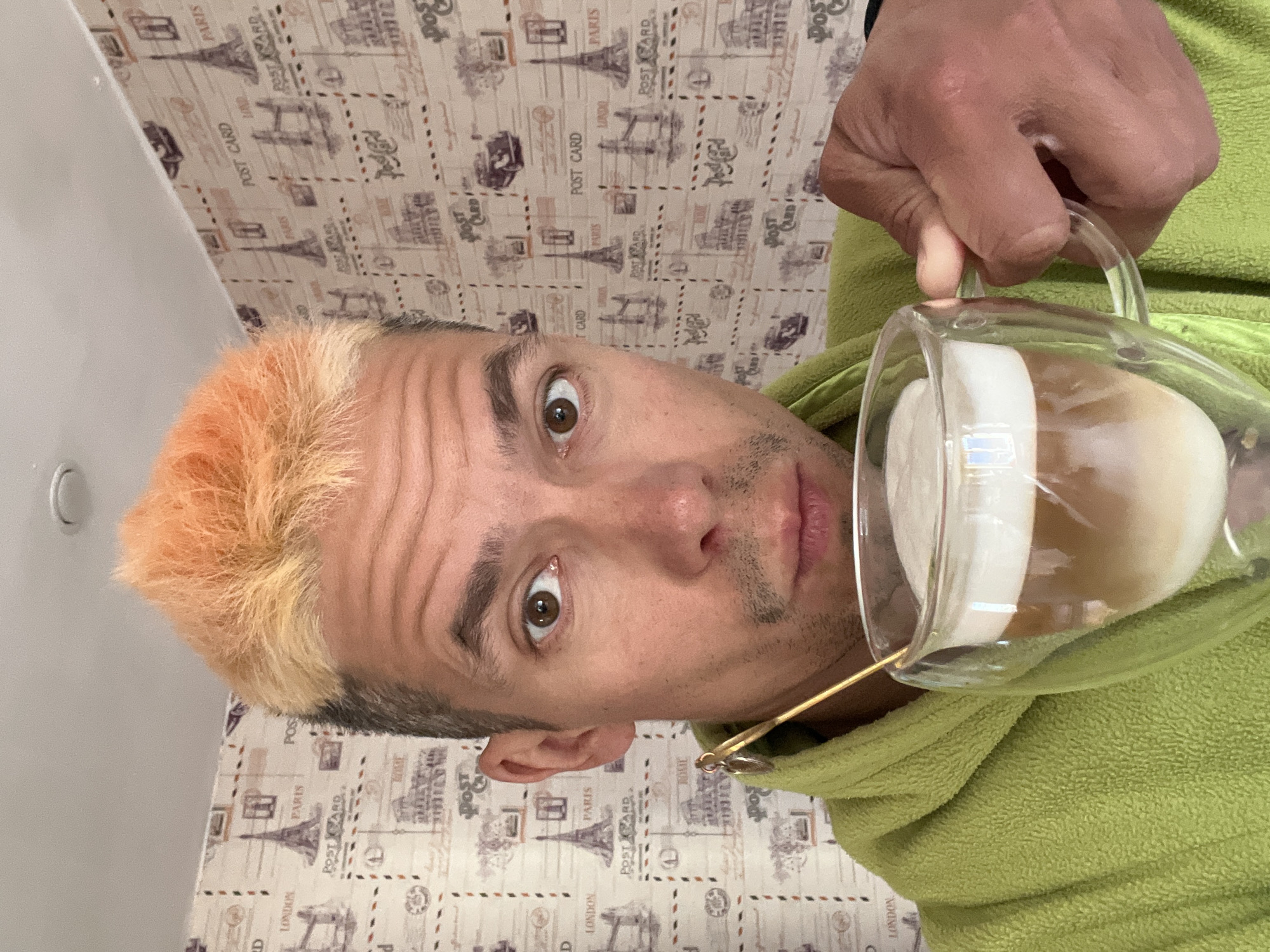 A man with two-toned orange and gray hair is holding a glass pitcher of coffee with milk or cream. He is wearing a green terrycloth bathrobe. The background is a patterned wallpaper with images of the Eiffel Tower and the words "Paris" and "London." He is looking into the camera. The image appears to be taken indoors with artificial lighting. I am unable to provide a location in the city of Chaikovsky, Russia.