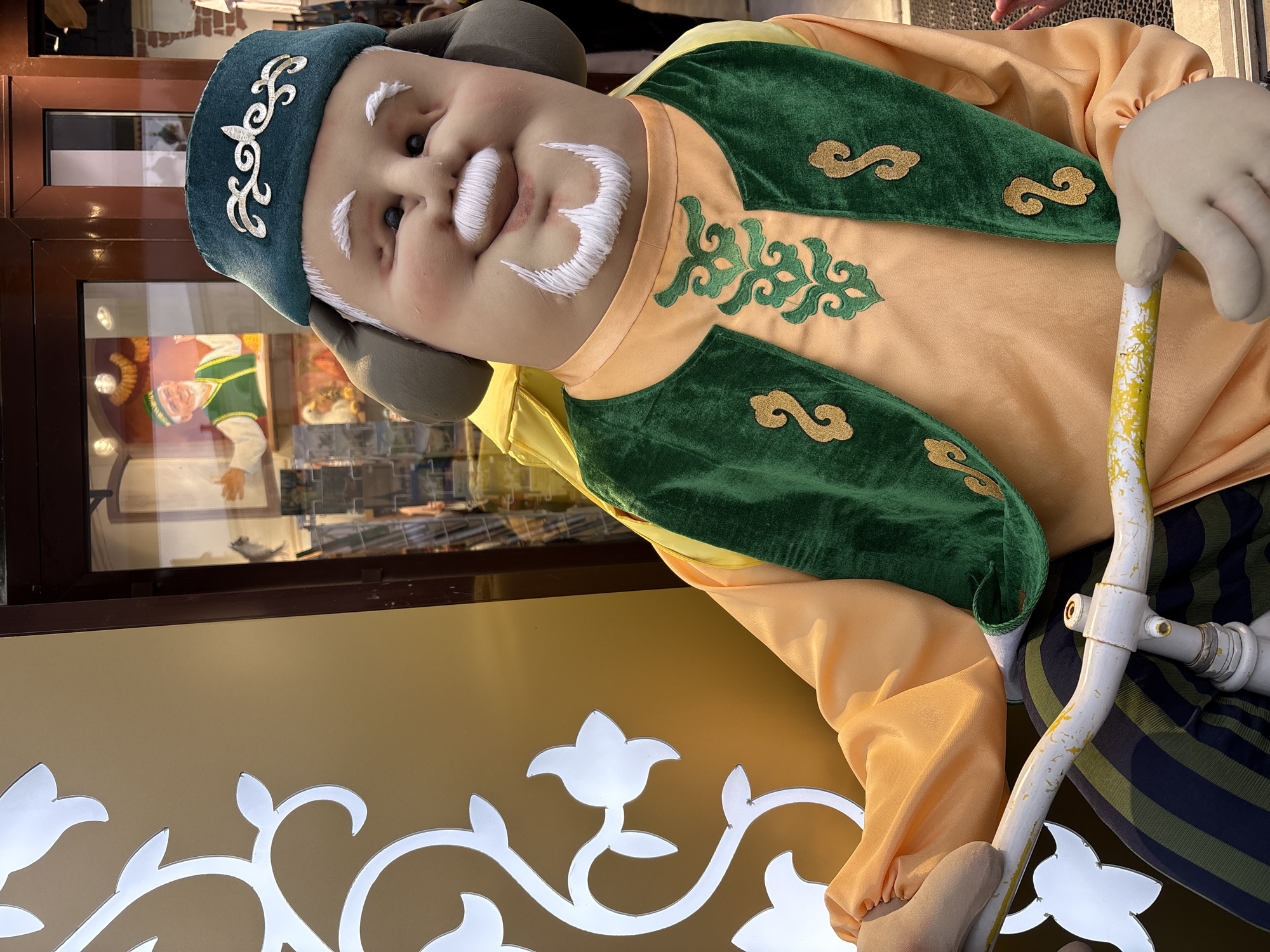 A large, cheerful-looking doll or mannequin, possibly representing an elder from a Turkic or Central Asian culture, is depicted as an outdoor display in Kazan, Russia. The figure wears traditional attire: a teal-colored cap featuring an ornate silver embroidered motif, a yellow-orange long-sleeved tunic, and a green velvet vest with decorative gold embroidery in swirling patterns. Its face displays a wide, jovial smile, with prominent white embroidered eyebrows, mustache, and beard. The doll's light-colored hand clasps a white cane with yellow distressed markings.
In the background, a large window reflects an indoor setting, including what appears to be another, smaller figure also dressed in green attire. The setting suggests the entrance area of a shop or restaurant. The foreground features a white, intricately cut metal or plastic panel with a flowing floral design. The scene is captured in daylight. The embroidery on the cap appears to be a traditional ornamental design rather than readable text.