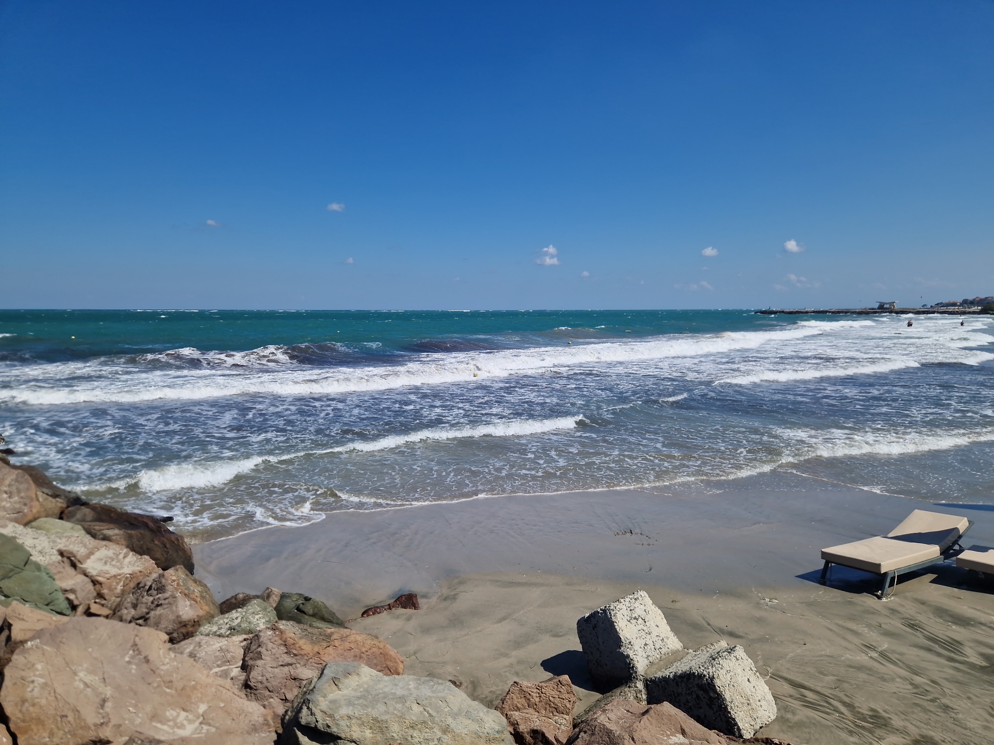 This image captures a scenic view of the beach in Pomorie, Bulgaria, on a clear, sunny day. The foreground is dominated by a rocky embankment on the left, composed of various-sized rocks in earthy tones, some with a greenish hue. In the middle ground, a sandy beach stretches out, with two light-colored lounge chairs positioned on the sand, one fully visible and the other partially. Several concrete blocks are scattered on the sand near the rocks and lounge chairs. The sea occupies the majority of the frame, with gentle waves breaking on the shore. The water exhibits a deep blue-green color. In the distance, a pier extends into the sea, with a small building visible at its end, and a few indistinct figures can be seen on or near the pier. The sky is a vibrant, cloudless blue, indicating good weather. The overall scene suggests a peaceful, coastal environment, likely a popular spot for relaxation by the sea.