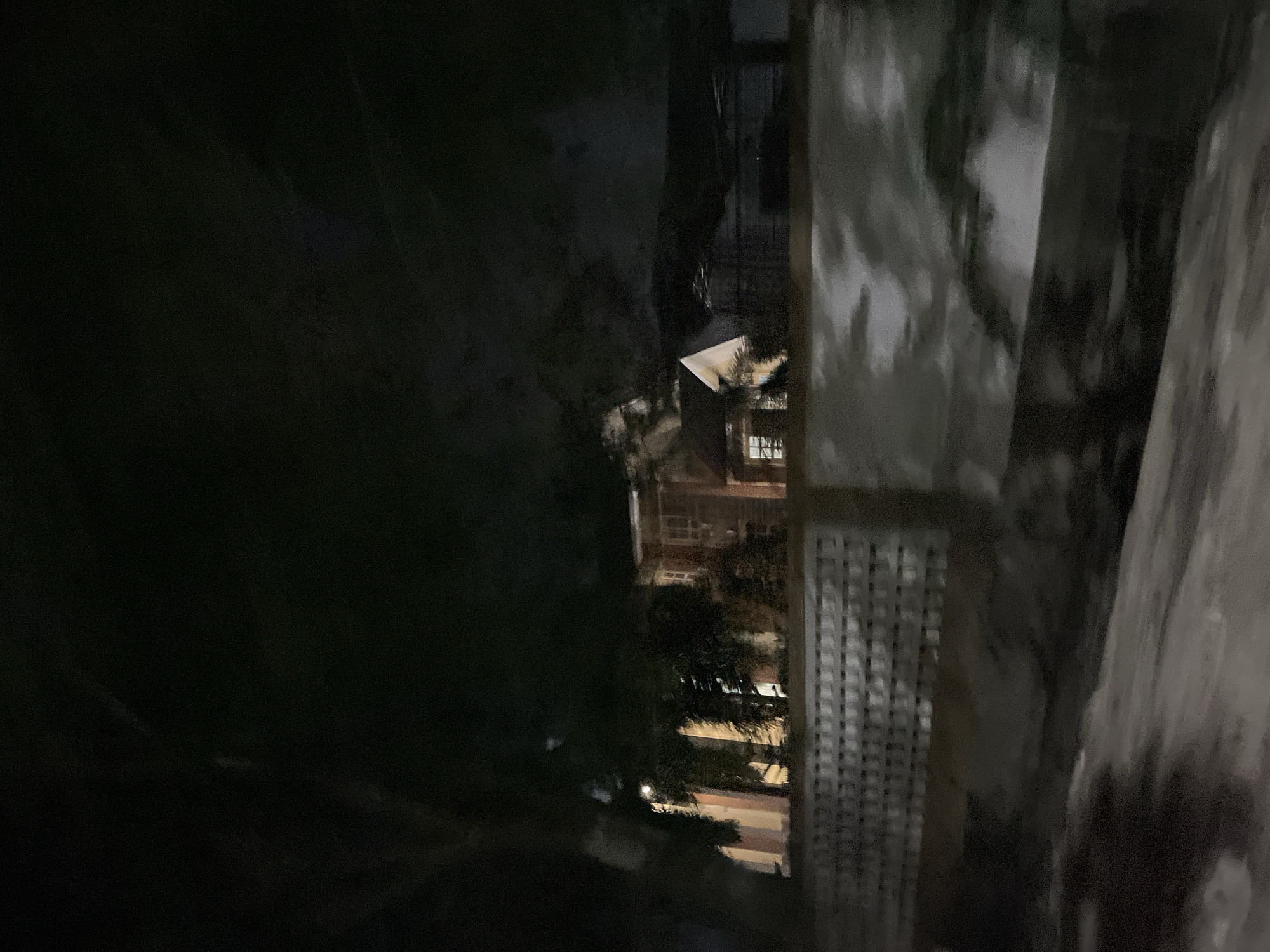 A dimly lit, outdoor nighttime scene captures a glimpse of a building structure in Bundar, Indonesia. The building appears to be a house or a public building with distinct architectural features, including multiple windows and what looks like a porch or balcony area. The structure is partially obscured by trees and foliage, adding to the moody, shadowed atmosphere. In the foreground, a patterned fence runs horizontally across the image, suggesting a separation between the viewer's vantage point and the property. The overall impression is one of a quiet, possibly residential area at night, with the faint glow from interior lights offering a subtle contrast to the darkness. The image's quality is somewhat grainy, likely due to the low light conditions and the capturing device.