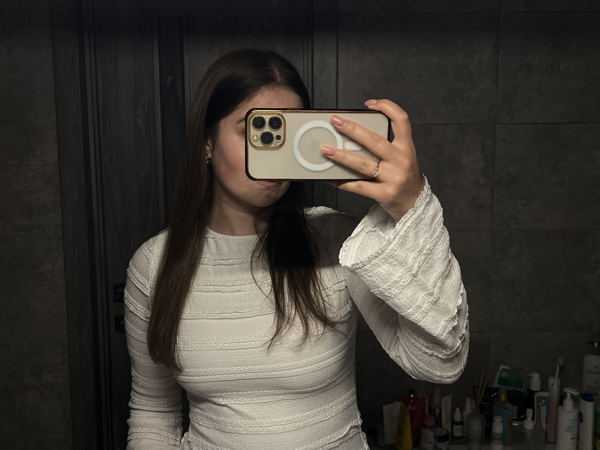 A woman is captured taking a mirror selfie in a dimly lit bathroom, likely in Ternopil, Ukraine. Her face is obscured by the smartphone she holds horizontally in front of her. She has long, dark brown hair that falls over her shoulders. She is wearing a white, long-sleeved top or dress with a textured, ribbed pattern and ruffled cuffs on the sleeves.
The smartphone appears to be an iPhone with a triple camera system, encased in a clear cover with a visible white circular MagSafe ring on the back. The phone itself has a gold or beige tone with a dark reddish-brown rim on the case. Her right hand, holding the phone, displays neatly manicured light pink nails and a ring with a clear stone on her ring finger. A small gold hoop earring is visible on her left ear.
The background consists of dark gray or black tiled walls, suggesting a modern bathroom aesthetic. To the right of the frame, a bathroom counter is visible, cluttered with an assortment of personal care products, including various bottles of lotions, shampoos, and a toothbrush, indicating a lived-in space. The low lighting suggests the photo was taken during the evening or night. No discernible text is visible on any objects in the image.