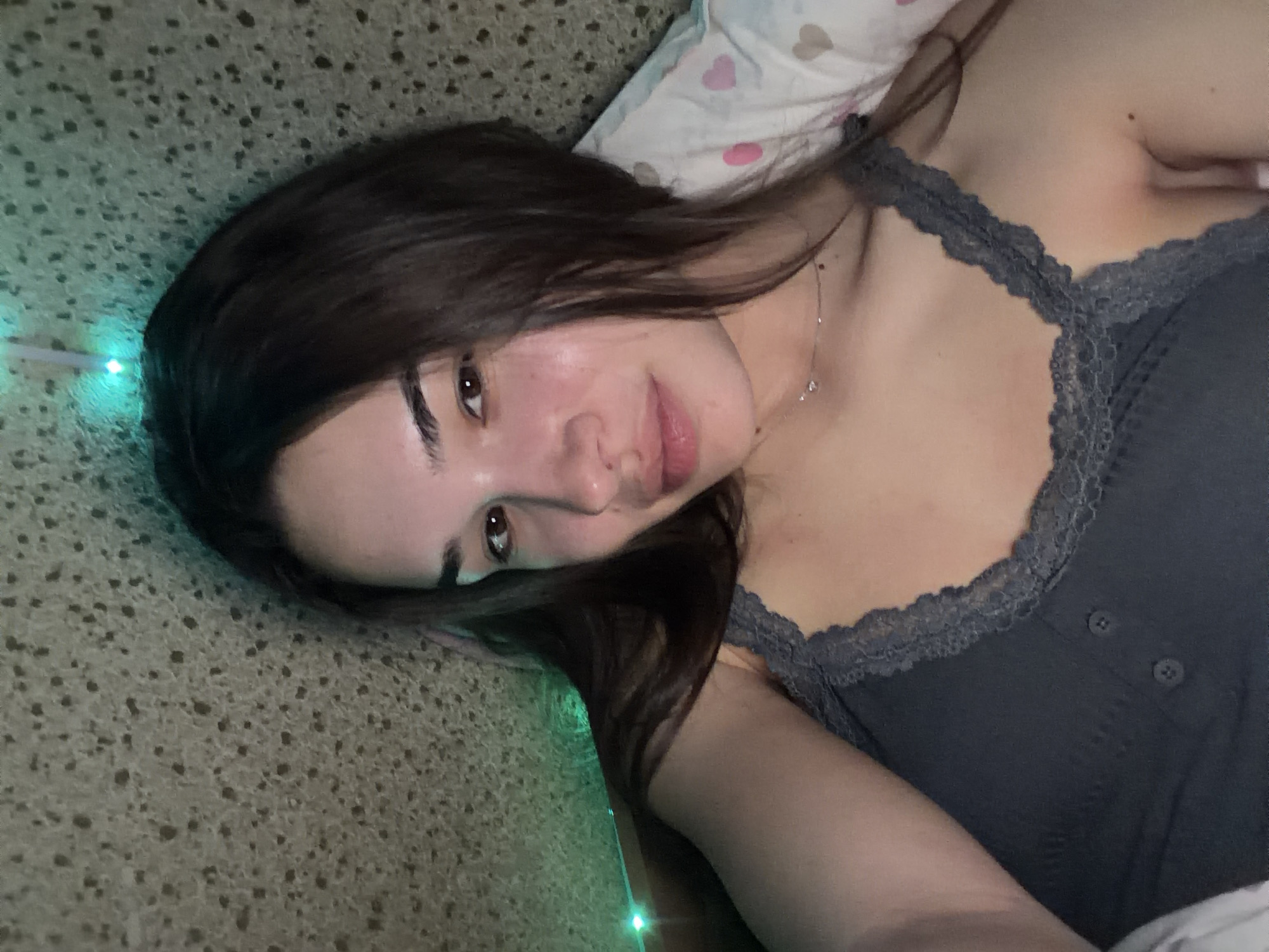 A young woman lies on her left side on what appears to be a textured surface, possibly a carpet or rug. She has long dark brown hair and is wearing a gray tank top with lace detailing. Her skin is light in color. A thin silver necklace is visible on her neck. Above her head is a white pillow with pink heart designs. In the background, there are blurred green lights. There is no verifiable information in the image to indicate a location in Sofia, Bulgaria.