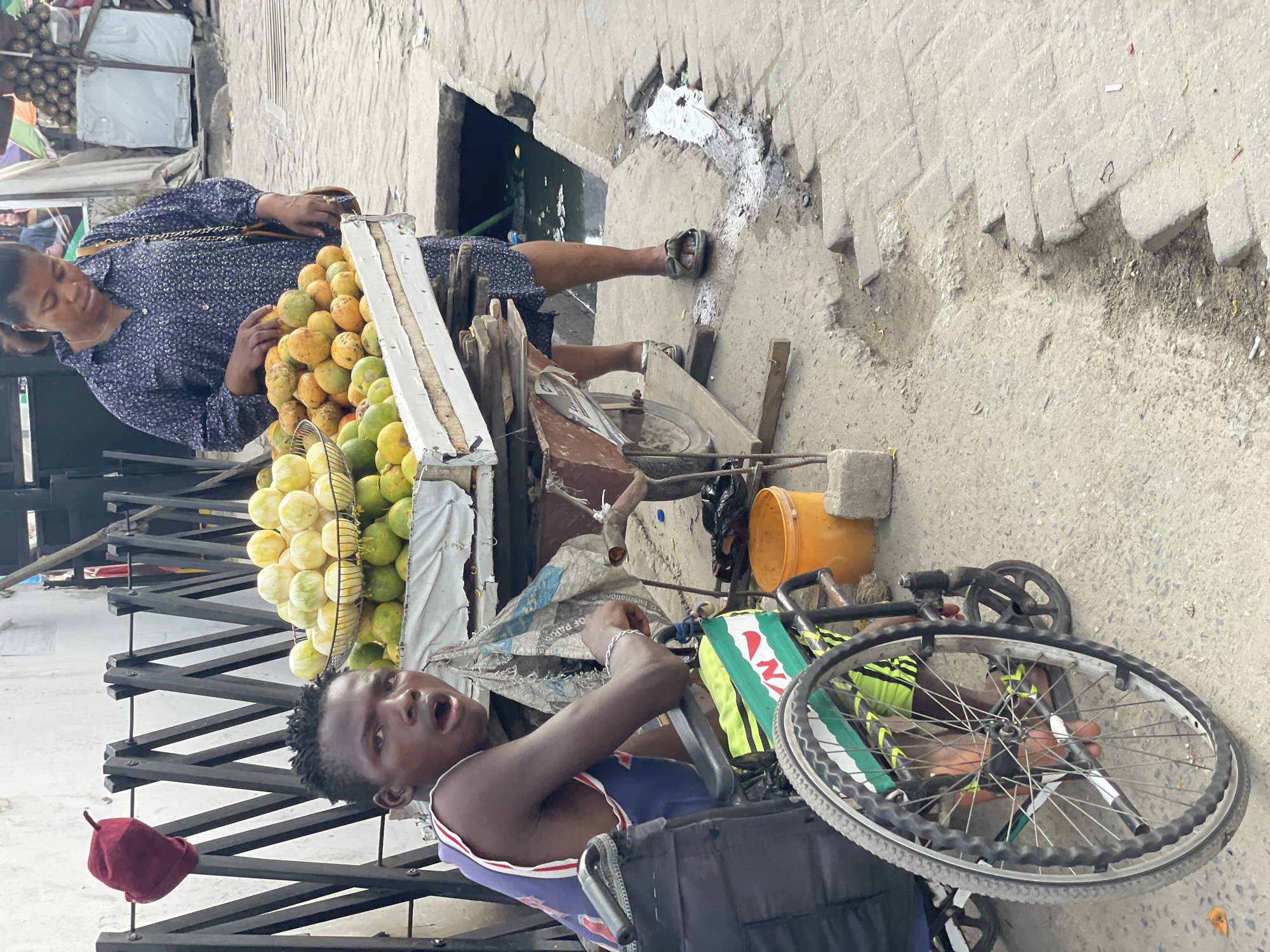 This street scene, likely in an informal market area in Ado, Nigeria, depicts daily life during the daytime. The ground is made of rough, uneven concrete pavers, with visible cracks and a section featuring a dark, watery drain or puddle.
In the foreground, a young man, possibly a vendor, is seated in a modified mobility device resembling a wheelchair or a hand-powered tricycle. He wears a blue and white striped tank top and has short, dark hair. His mouth is slightly open, and he looks directly towards the camera, conveying an open or surprised expression. A red, traditional cap rests on a metal barrier to his left. His mobility device has a green and white banner on its side, displaying fragmented text including "OF PARKS" and a partial "N".
To the young man's right, an adult woman stands next to a fruit cart, which is heavily laden with an abundance of yellow and green fruits, likely mangoes or guavas, piled high on a display stand. She is dressed in a dark blue, long-sleeved patterned dress and wears sandals. Her attention is focused on the fruit, suggesting she is in the process of selecting items or making a purchase. She carries a bag with a metallic chain strap over her shoulder.
The scene captures an everyday interaction between a vendor and a customer, set against a backdrop of urban infrastructure and informal commerce under bright, clear daylight conditions.