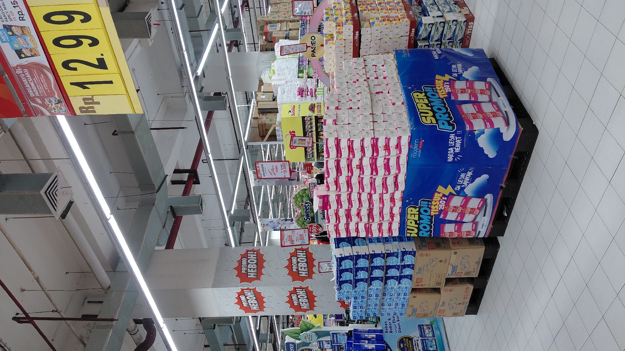 This is an eye-level, medium shot of a supermarket aisle in Purwokerto, Indonesia, featuring promotional displays of facial tissues and snacks. The aisle is brightly lit by fluorescent ceiling lights. In the foreground, large stacks of white and pink packaged facial tissues are arranged on blue and black pallets, with prominent "SUPER PROMO" signage. To the left, stacks of blue packaged items, likely diapers or sanitary products, are also on display. Further back, shelves are stocked with various grocery items, including snacks like "ATOM" brand chips. A large yellow price sign prominently displays "Rp. 12.99" along with "DIRGAHAYU 81 INDONESIAKU 17 AGUSTUS 2020". The floor is tiled in a grid pattern. There are no identifiable people in the immediate foreground or mid-ground, though blurred figures may be present in the background. The overall scene depicts a commercial retail environment during a promotional period.