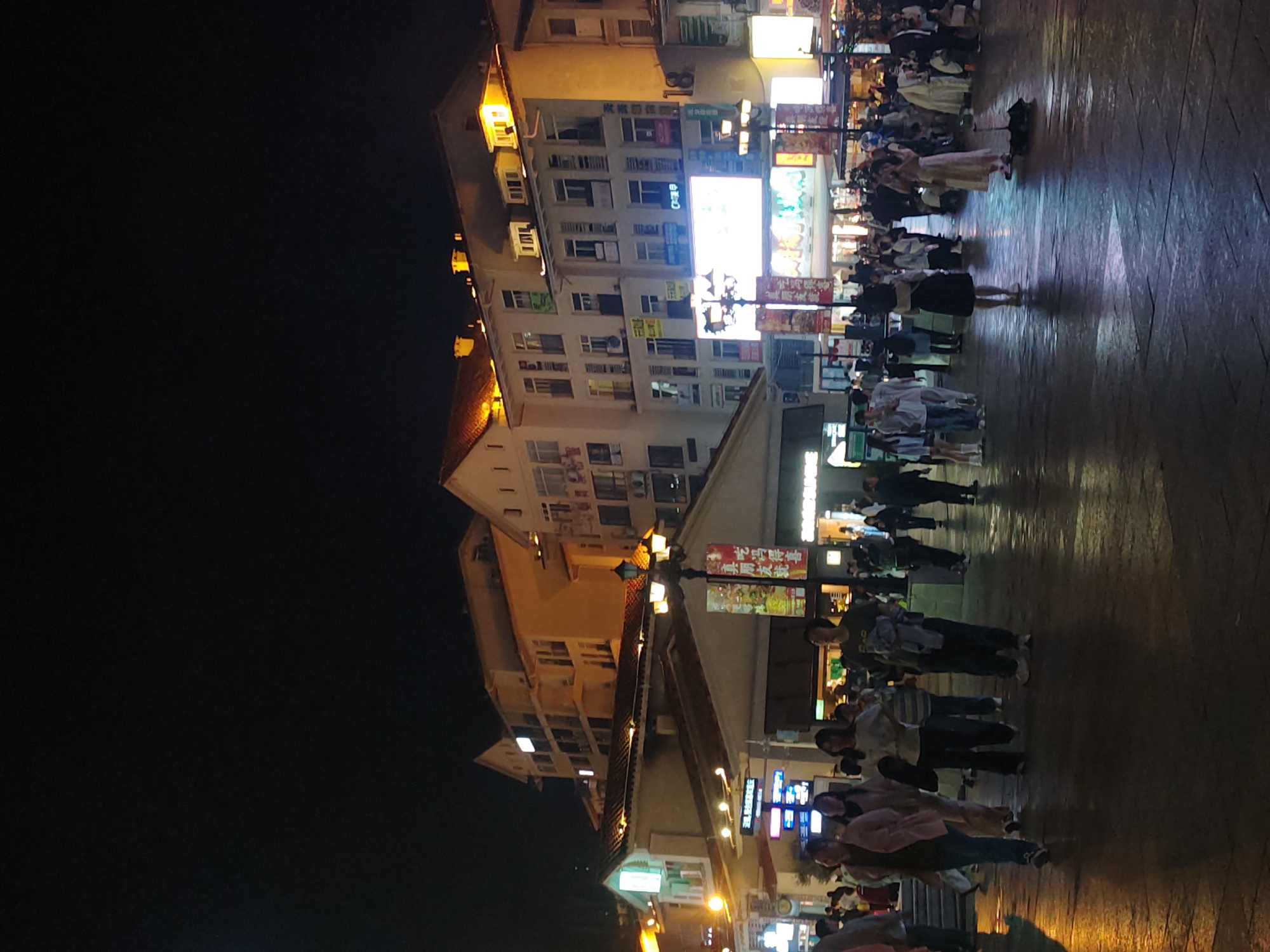 The image depicts a nighttime scene in Chongqing High-tech Zone, China. Illuminated buildings line a pedestrian area, where numerous people are walking. Light posts and building signs illuminate the scene. The ground is paved with large stones reflecting the artificial light. The sky is dark.