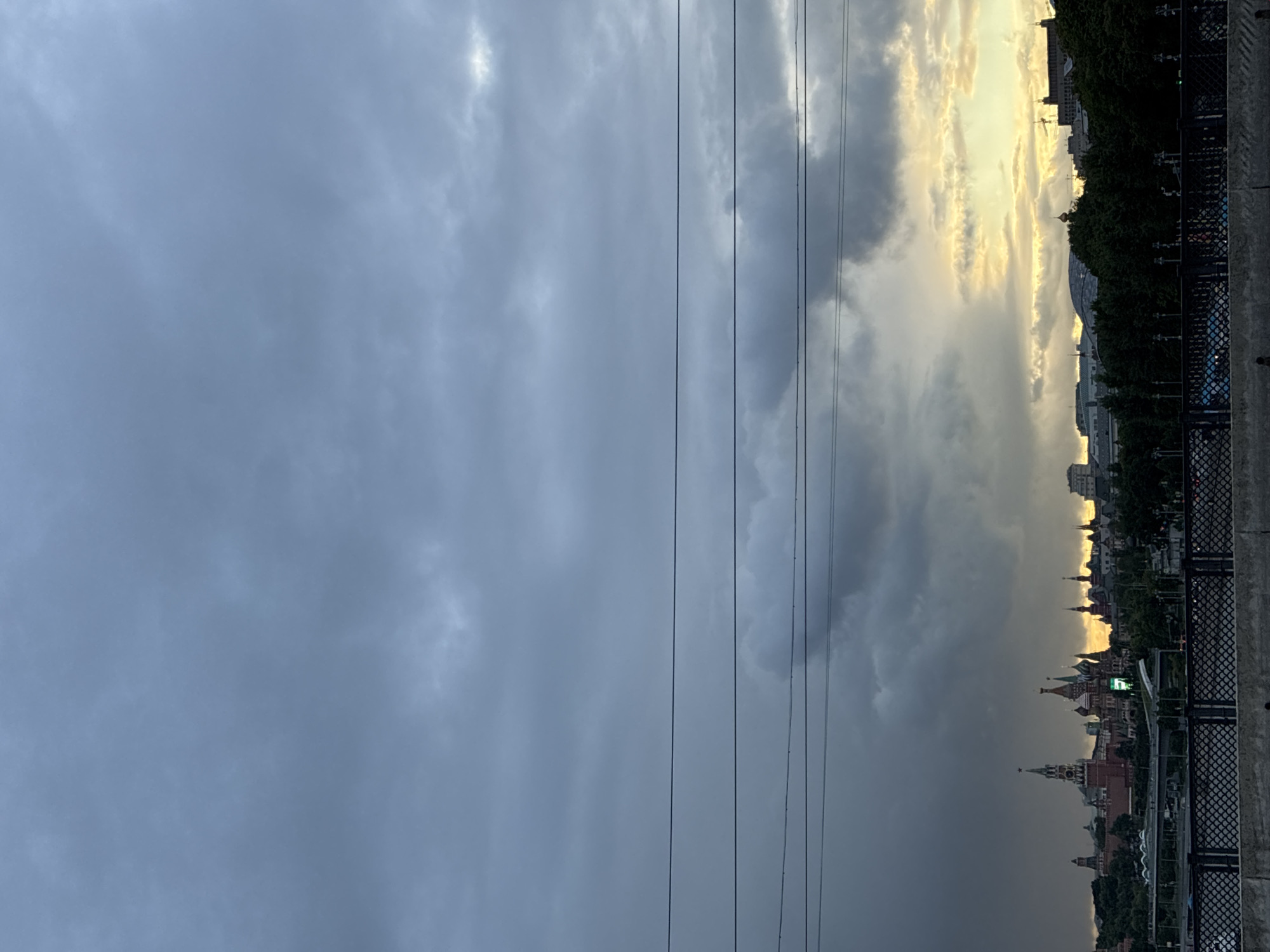 The image shows a view of Moscow, Russia under a cloudy sky. Power lines stretch vertically in the center of the frame. To the right, the Moscow Kremlin and other buildings are visible in the distance, illuminated by a bright light source. The right edge of the frame shows a concrete wall or embankment.