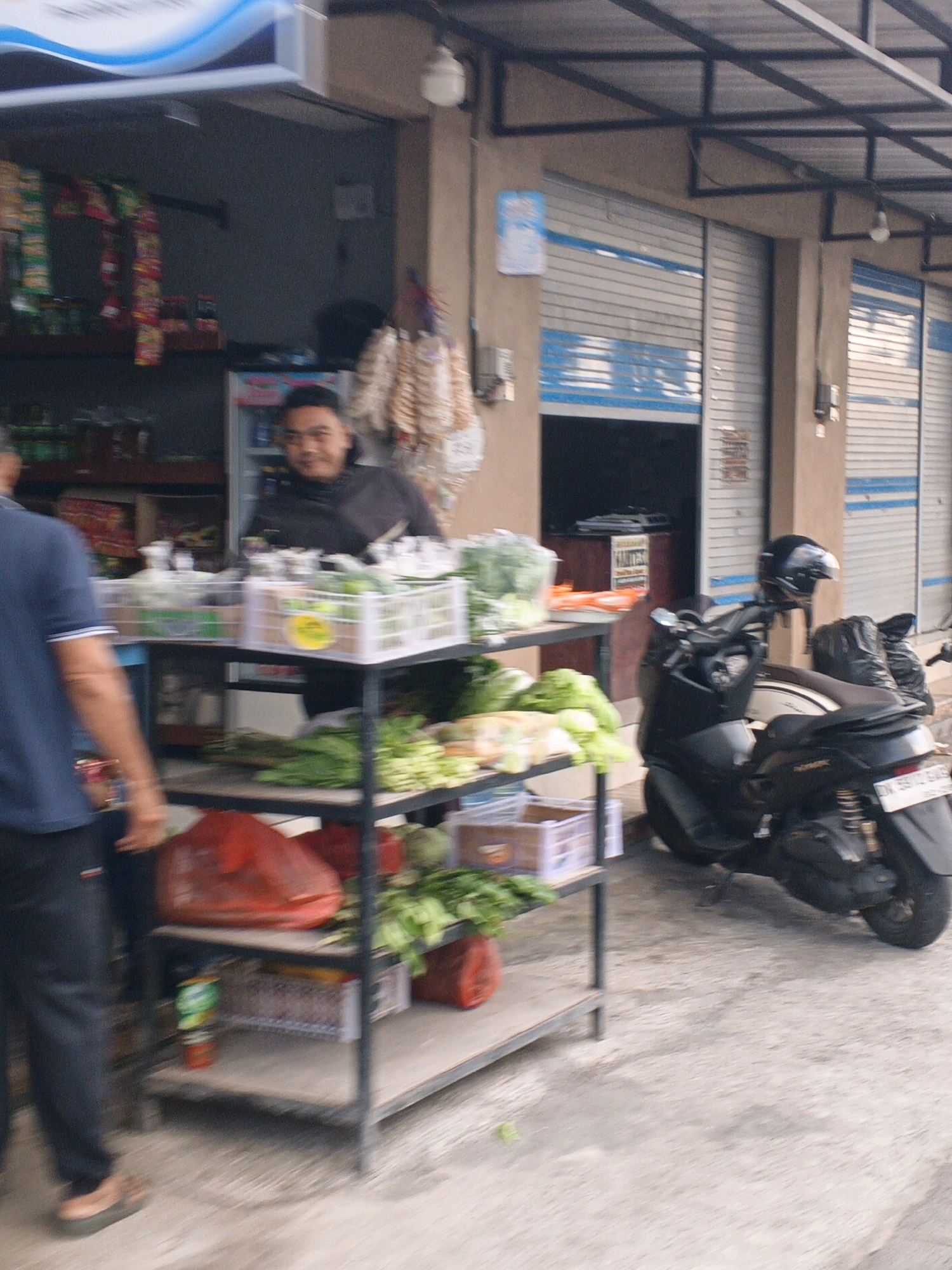 This is a medium shot of a street market stall in Denpasar, Indonesia. A man is standing behind a display of fresh produce, including leafy green vegetables and what appear to be cucumbers or similar items. The stall has multiple shelves stocked with various items, including bagged snacks on the upper shelves and what seem to be plastic containers of produce on the middle shelves. A red plastic bag and other items are visible on the lower shelves. To the right of the stall, a black motorcycle is parked, with a black helmet resting on the handlebars and a black bag secured to the rear. The background shows the facade of a building with closed roller shutters, typical of many shops in the area. The ground is paved with concrete. A person in a blue shirt and dark pants is partially visible on the left side of the frame, walking away from the camera. The scene depicts a typical day at a local market.