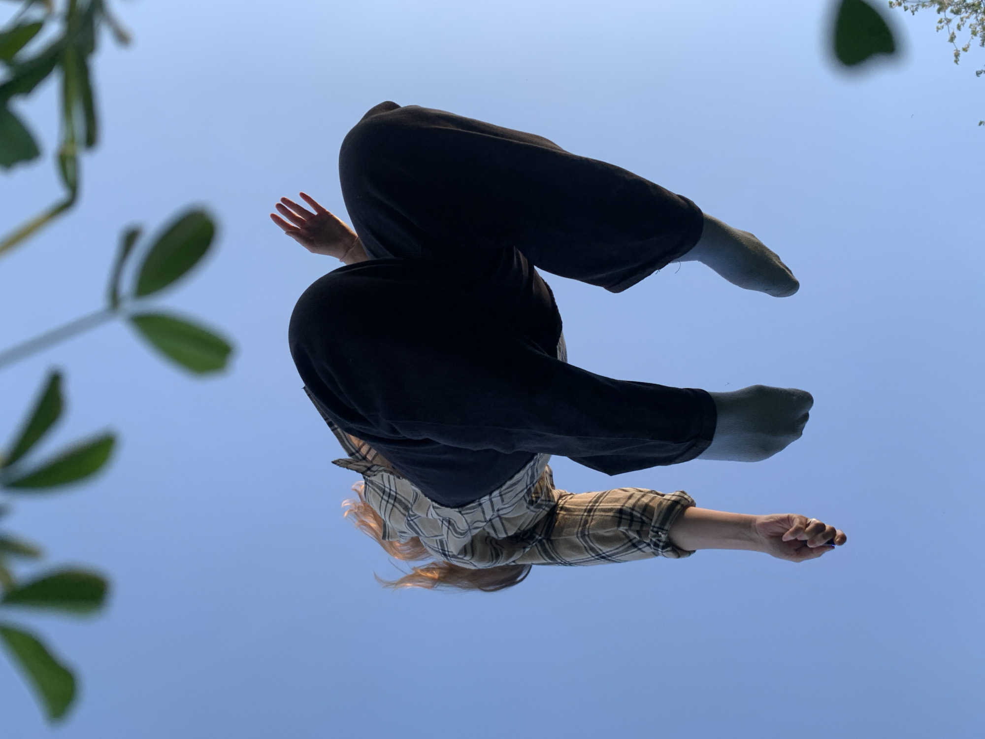 A low-angle shot looking up at a person, likely standing on a glass surface or lying on their back, against a clear blue sky. The person is wearing black pants, grey socks, and a plaid shirt. Their arms are extended, one hand open and the other holding a blue object. Some blurred greenery is visible in the periphery of the frame. The location is purportedly Chaikovsky, Russia.