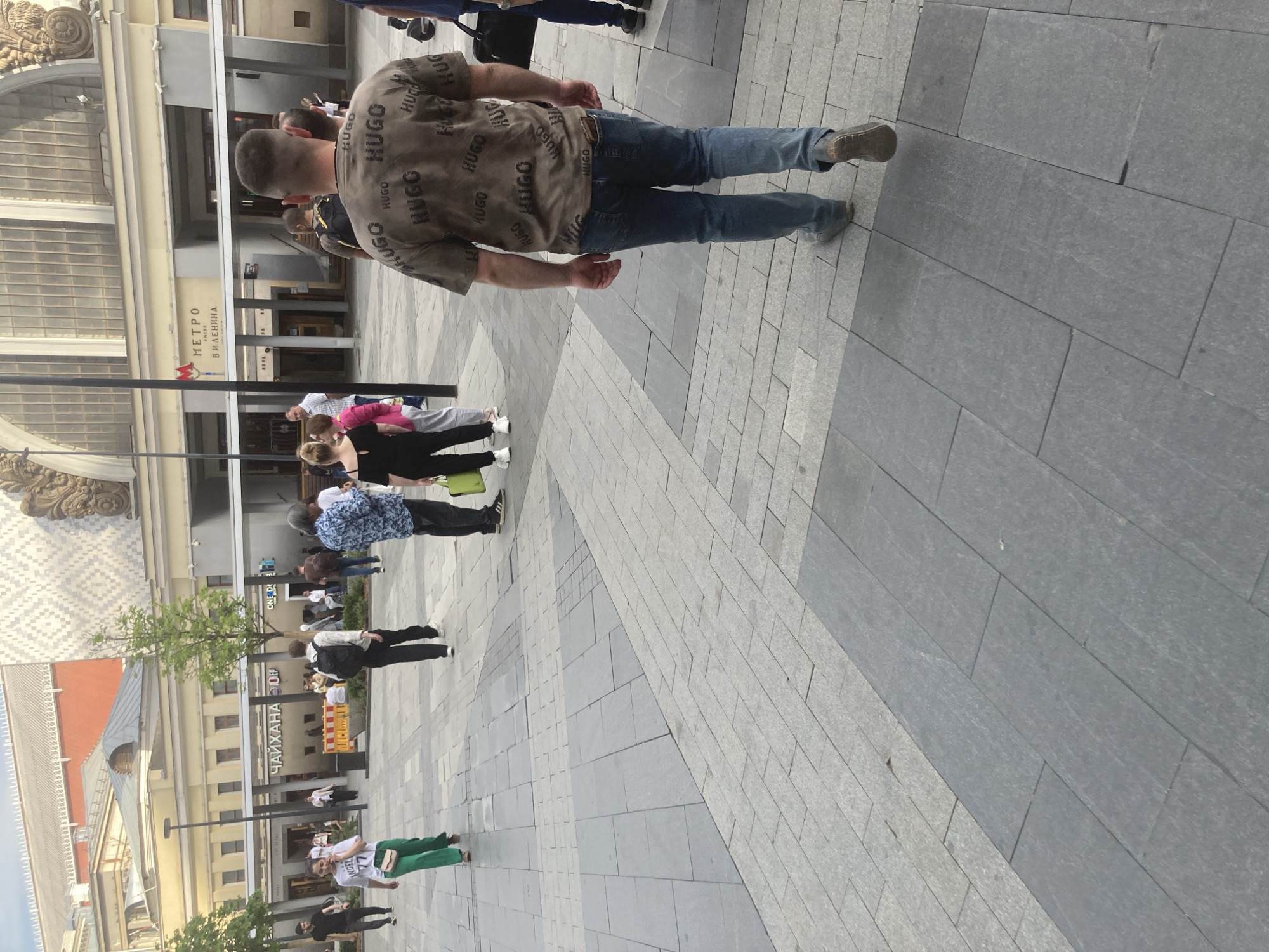 The image shows a pedestrian area outside what appears to be a metro station in Moscow, Russia. The station, identified by a sign with the word "METPO" and "В И ЛЕНИНА" (V.I. Lenin's) in Cyrillic, indicates that the location is likely near a metro station named after Lenin. A man wearing a t-shirt covered with the word "HUGO" and jeans is walking away from the camera. Several other people are also present, some standing in groups and others walking in different directions. There are several storefronts visible, including a restaurant named "ЧАЙХАНА ОЧЕР" (Chaikhana Ocher) and a business called "ONE De". The environment includes paved walkways with varying patterns, buildings with architectural details, and some greenery.