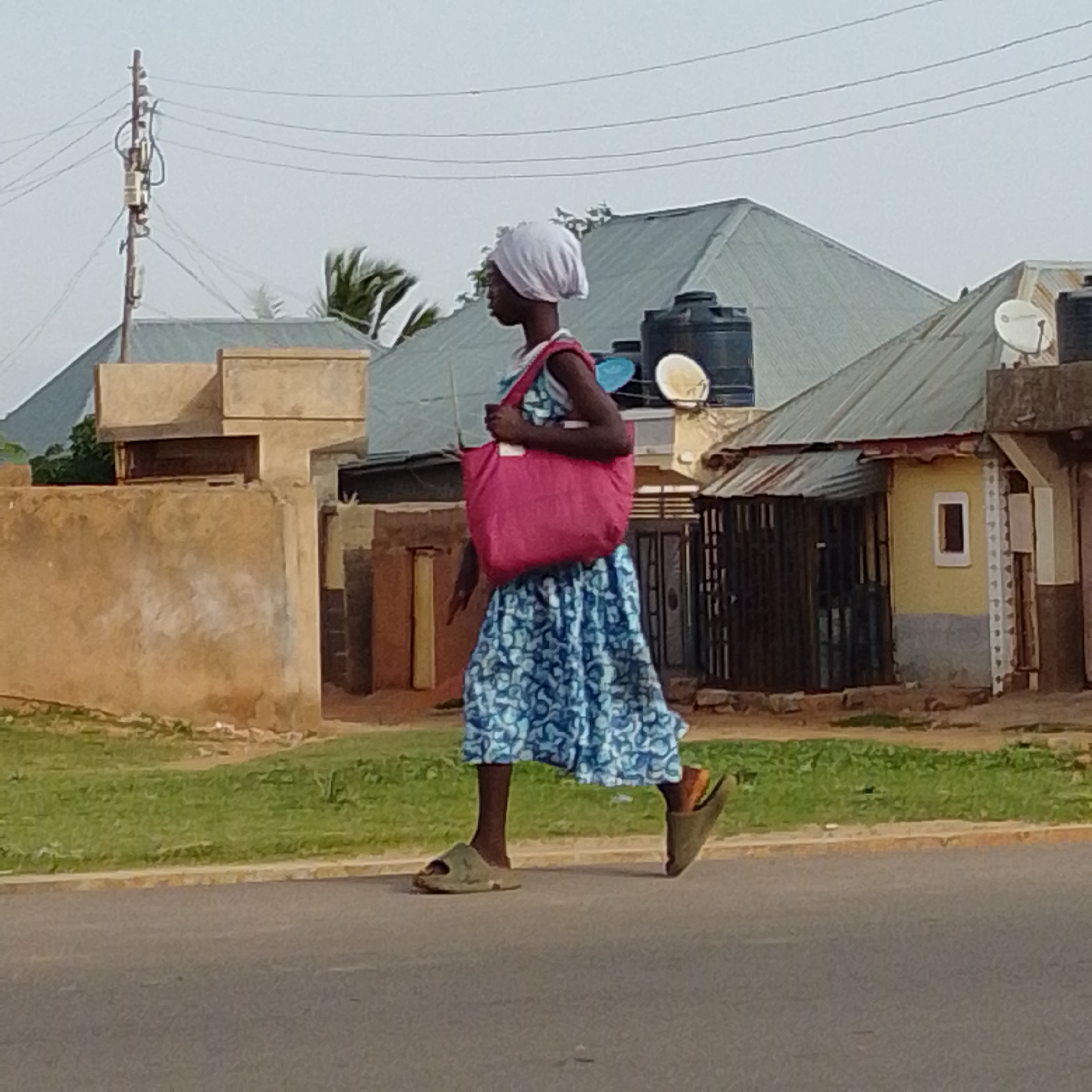 The image depicts a young girl walking on a paved road in Gombe, Nigeria, carrying a large pink bag slung over her shoulder. She is wearing a white headscarf and a blue and white patterned dress. Her feet are clad in olive green slides. The background shows a residential area with several buildings, some with corrugated metal roofs. A utility pole with electrical wires is visible on the left side of the image, and a large black water tank is on the roof of one of the buildings. The ground is covered with green grass along the roadside. The overall scene suggests a typical day in this Nigerian city.