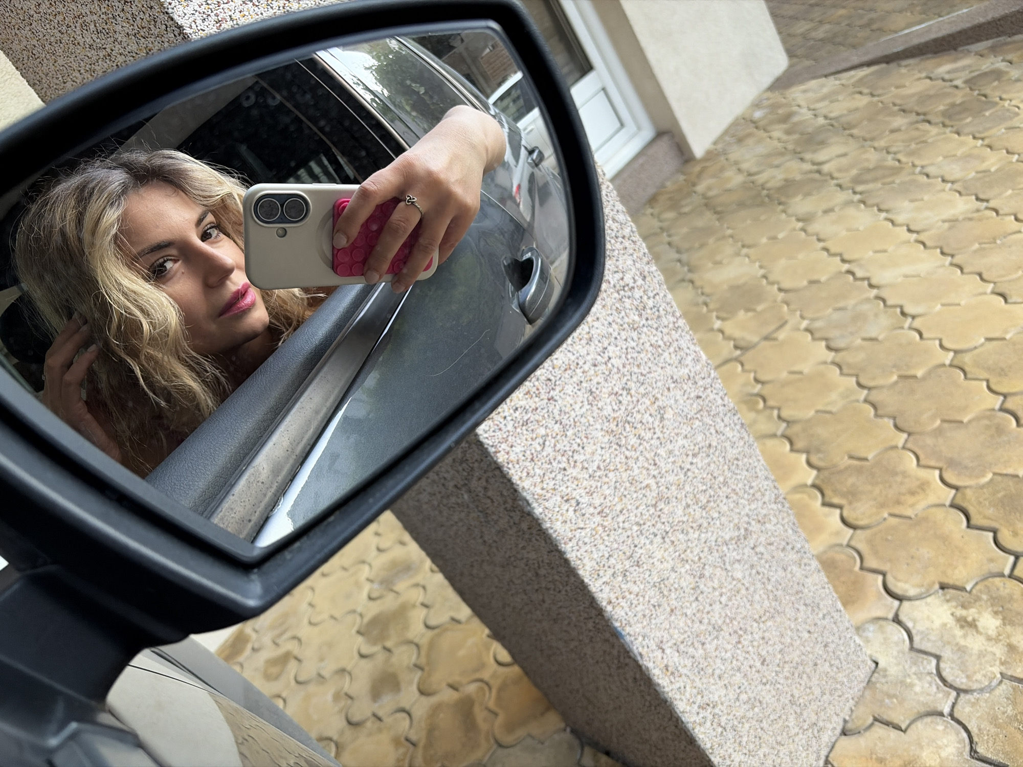 The image shows a selfie taken in a car's side mirror, reflecting a woman with blonde wavy hair, holding a white smartphone with a pink grip. The background visible in the mirror includes the interior of the car and a glimpse of a building exterior. The surrounding environment outside the car consists of a patterned stone pavement and a textured pillar or wall. While precise location data is unavailable, the request indicated Budva, Montenegro.