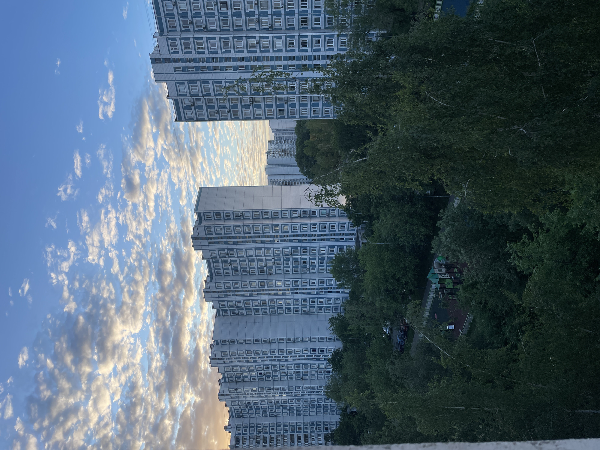 The image shows a high-angle view of a Moscow residential neighborhood at dusk. The sky on the left side of the frame is filled with altocumulus clouds illuminated by the setting sun, transitioning from blue to shades of gold and pink. Several multi-story residential apartment buildings dominate the scene, with a repetitive grid of windows indicating a standardized, Soviet-era architectural style. Green trees obscure portions of the buildings and also cover the ground below, and include a playground with green and red elements. Several cars are parked in the shadows. The view is taken from an elevated position, possibly from another apartment building. The overall atmosphere is one of quiet urban life at the end of the day.