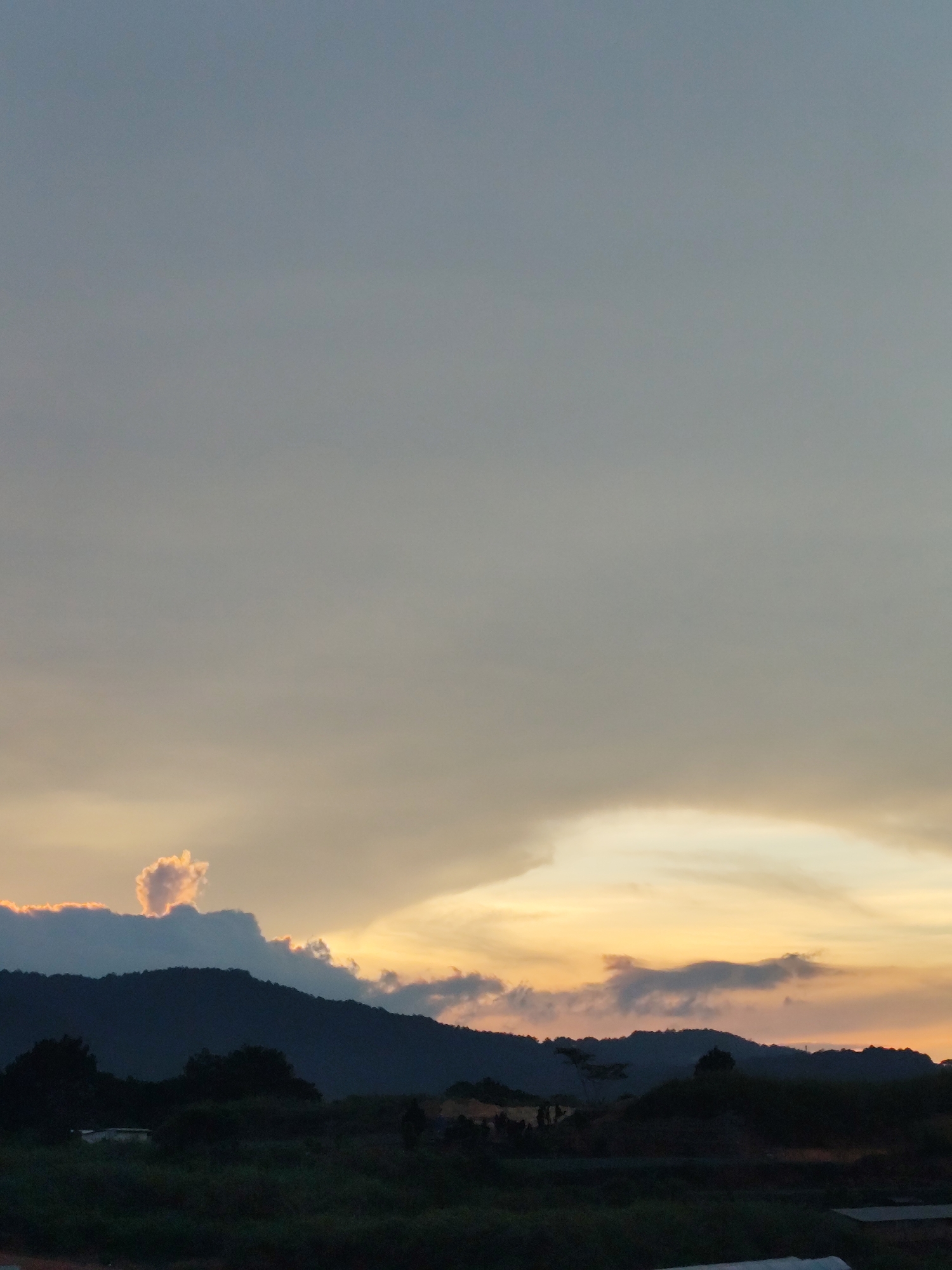 The image depicts a landscape at dusk, possibly in Da Lat, Vietnam, though no specific landmarks are clearly visible. A dark, silhouetted mountain range dominates the lower portion of the frame, with low-lying vegetation in the foreground. Above the mountains, the sky exhibits a gradient of colors associated with sunset, transitioning from a lighter golden hue near the horizon to a muted grey-blue higher up. There are clouds in the sunset sky, some with distinctive shapes and orange tinges. The scene has a calm, rural or semi-rural feel due to the expansive sky and natural landscape. No people are explicitly visible.