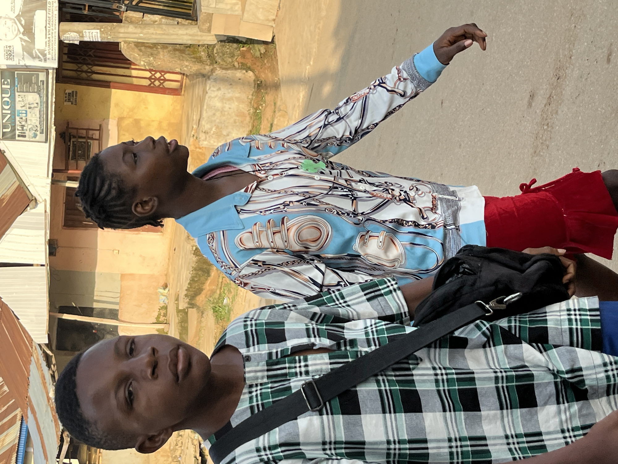 In Abuja, Nigeria, two young individuals are captured walking on a paved street during daylight. The person on the left, facing forward, wears a green, black, and white plaid shirt and a black crossbody bag. Their gaze is directed ahead, with a neutral expression. To the right, a second individual is seen in profile, walking forward and slightly to the right, with their head turned towards the viewer. They are dressed in a light blue collared shirt featuring a print of chains and buckles, paired with red shorts. The background reveals a row of buildings with corrugated metal roofs and plastered walls, characteristic of the urban landscape in Abuja. The lighting suggests it is either early morning or late afternoon, casting a warm glow on the scene.
