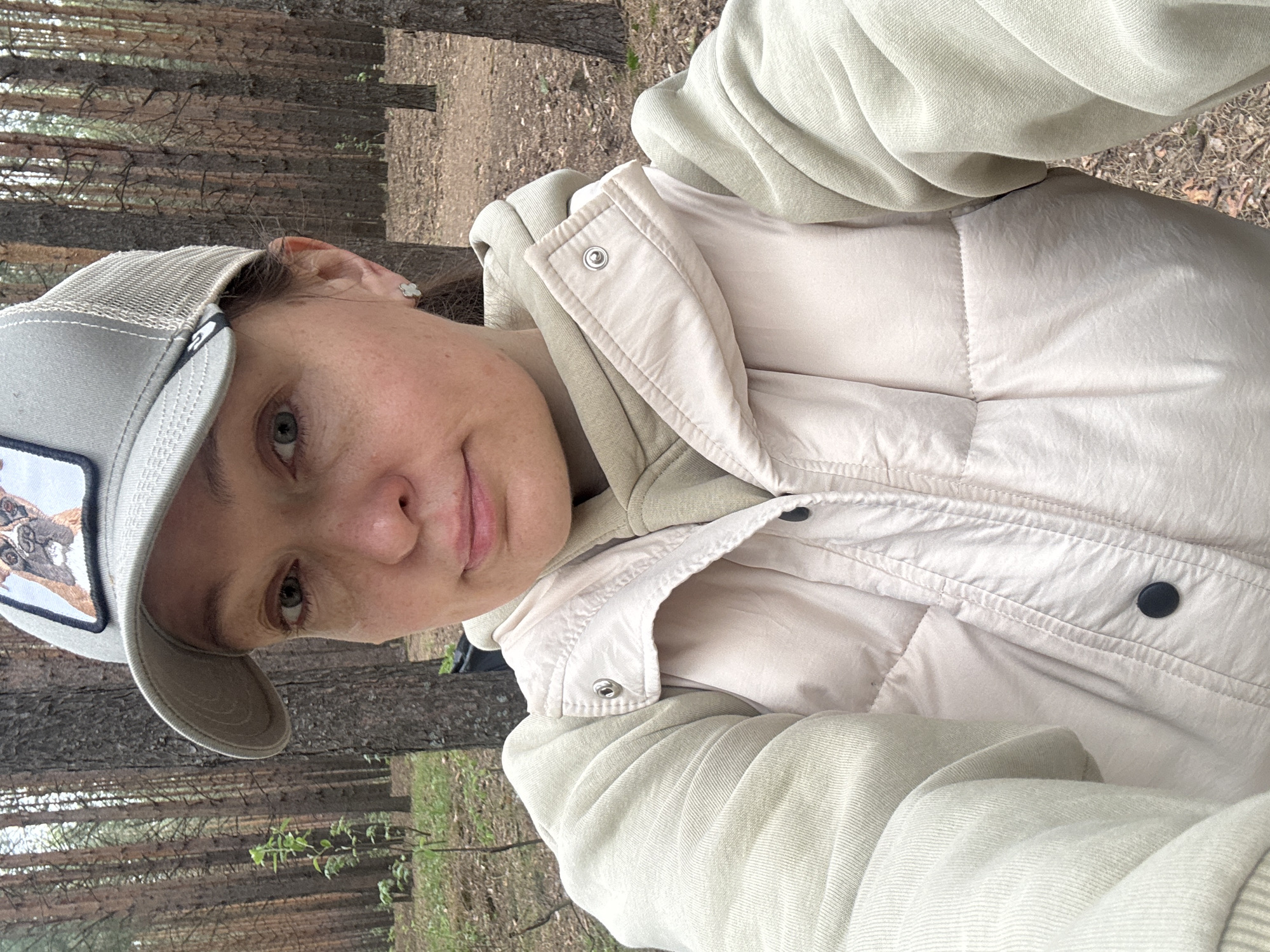 A woman in a beige cap with a dog patch, a beige hoodie, and a beige quilted vest takes a selfie in a pine forest. The ground is covered in fallen pine needles. The location is likely in Chaikovsky, Russia, given the prompt request.