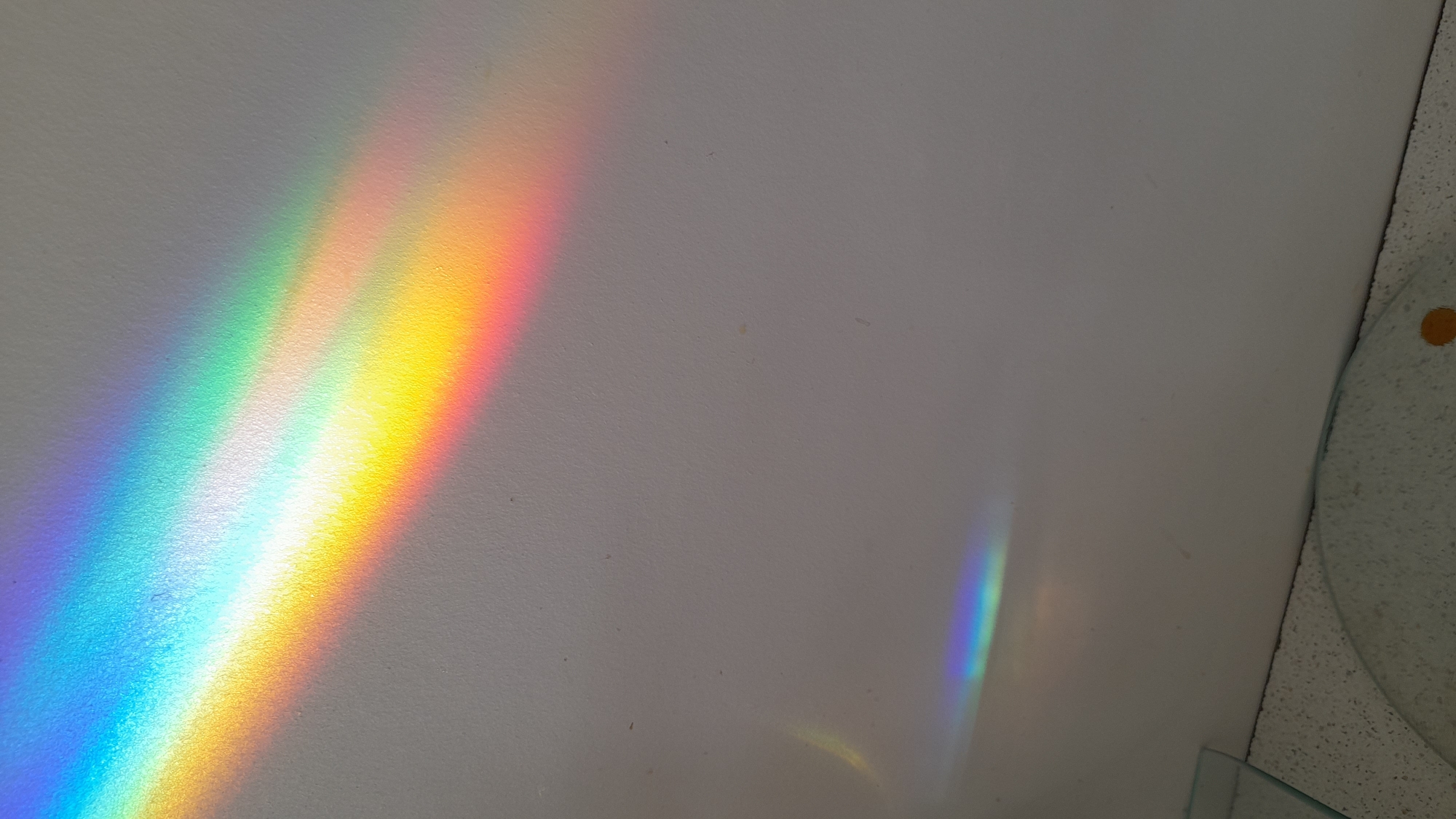 A rainbow spectrum of light is projected onto a white wall, displaying distinct bands of color from violet to red. A smaller, fainter rainbow is visible in the lower right quadrant of the image. The edge of a round, clear object with a textured surface is in the lower right corner of the photo. It is impossible to discern the location in the city of Saint Petersburg, Russia or information about people, actions, or interactions.