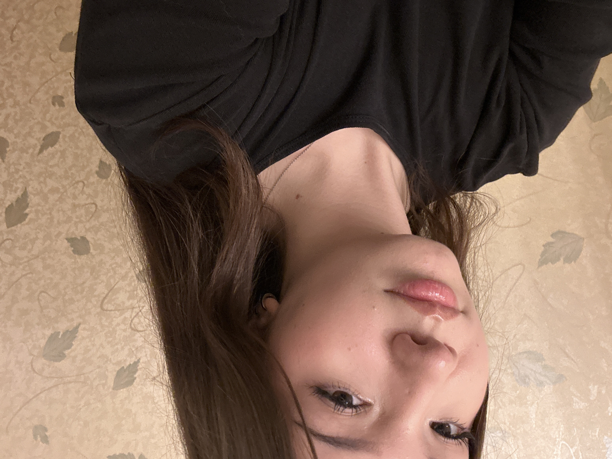 The image displays a head and upper torso portrait of a young individual, likely female, with light skin and long, straight dark brown hair. She is looking directly towards the camera with a neutral expression, her lips slightly parted and glossy. Visible facial features include dark brown eyes with defined lashes, dark eyebrows, and a small, shiny hoop earring in her left ear (viewer's right). A small, silver-colored chain necklace with a tiny, indistinct pendant is worn around her neck, where a small mole or blemish is also present. She is dressed in a plain black, long-sleeved, crew-neck top. The background consists of a light beige or cream-colored wallpaper patterned with repeating, muted grey-green stylized leaves and subtle swirling vine motifs. The lighting is even and appears to be indoor artificial illumination. The image does not contain any discernible visual elements to identify a specific geographic location, such as the city of Naberezhnye Chelny, Russia.