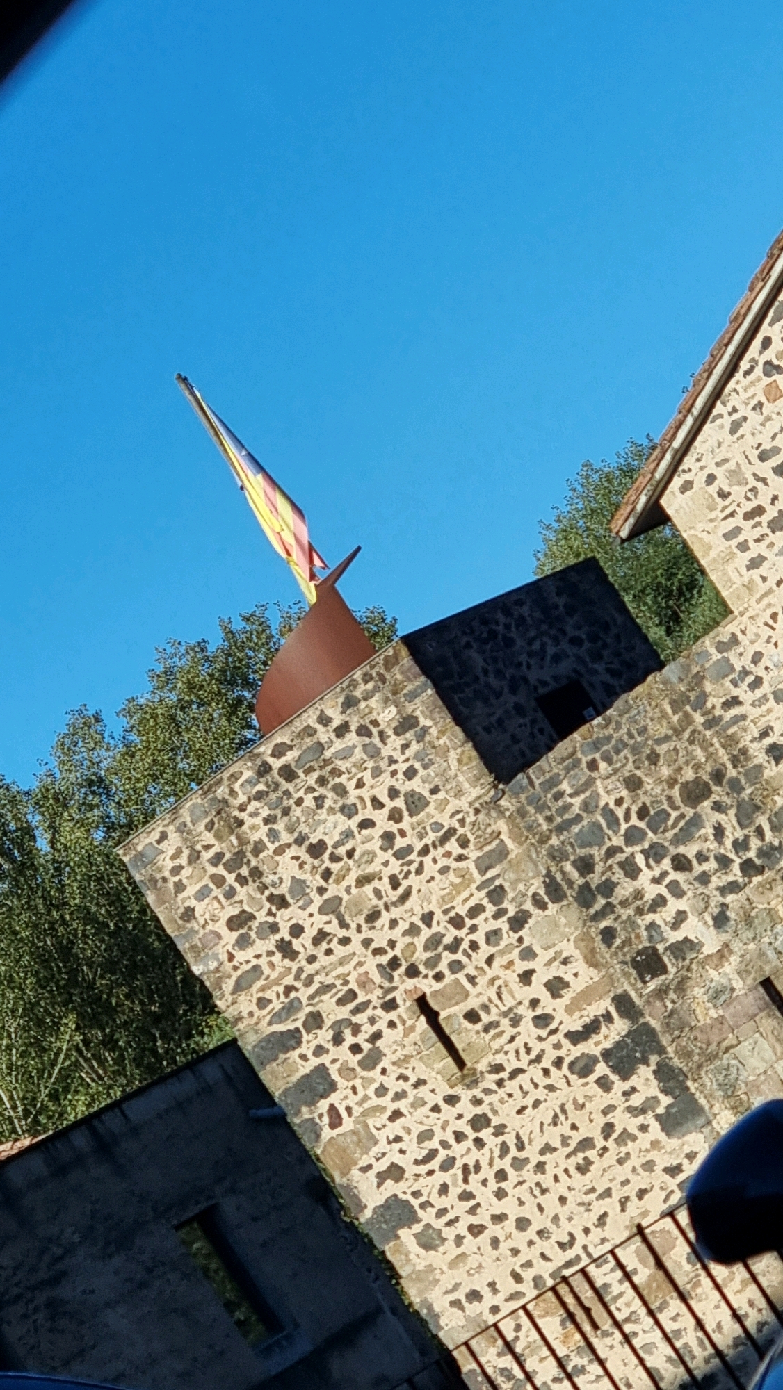 The image captures a view looking up at a stone building and a flag against a clear blue sky. The building appears to be made of rough, light-colored stones with some darker stone accents, suggesting a rustic or historical architectural style. A portion of a brown roof with a darker edge is visible at the top right. A dark, angular structure juts out from the stone building.
Dominating the upper part of the image is a flag with horizontal stripes in yellow and red, with a section of red and yellow bands visible at the top. The flag is attached to a thin pole and appears to be fluttering. Behind the building, lush green trees are visible.
While the image itself does not explicitly state its location, the flag's design is consistent with the Catalan flag, suggesting a location in Catalonia, Spain. Sant Joan Les Fonts is a municipality in the Garrotxa region of Girona, Catalonia, Spain. If this image were taken in Sant Joan Les Fonts, it would be depicting a scene within this town. The overall impression is of a bright, sunny day with a focus on the architectural elements and the flag. There are no people or discernible actions depicted in the image.