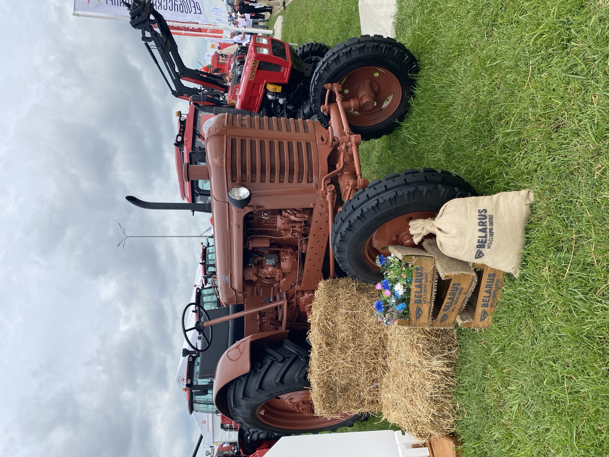 The image shows a brown tractor standing on grass in Voronezhskaya, Russia. The tractor is positioned next to a bale of hay with a crate containing flowers and a burlap sack labeled "BELARUS" on top. In the background are red tractors and some people. The sky is overcast.