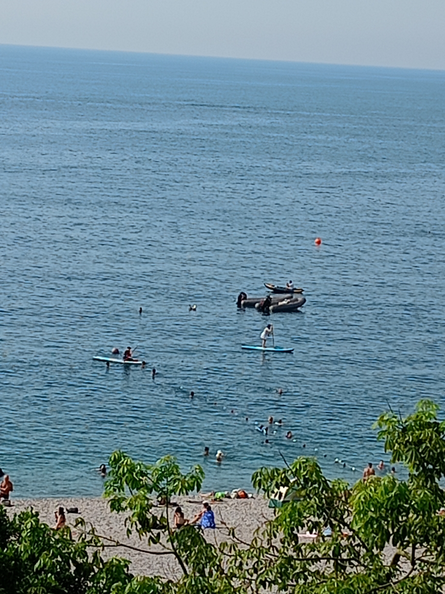 The image shows a view of the Mediterranean Sea from a beach in Antalya, Turkey. The water is a clear blue, with gentle waves. Several people are enjoying the water, with some swimming and others paddleboarding. There is a small inflatable boat with people in it, and another person on a paddleboard further out. The beach is sandy, with some rocks and vegetation along the shoreline. Trees with green leaves are in the foreground, obscuring some of the view of the beach. The sky is a pale blue and hazy.