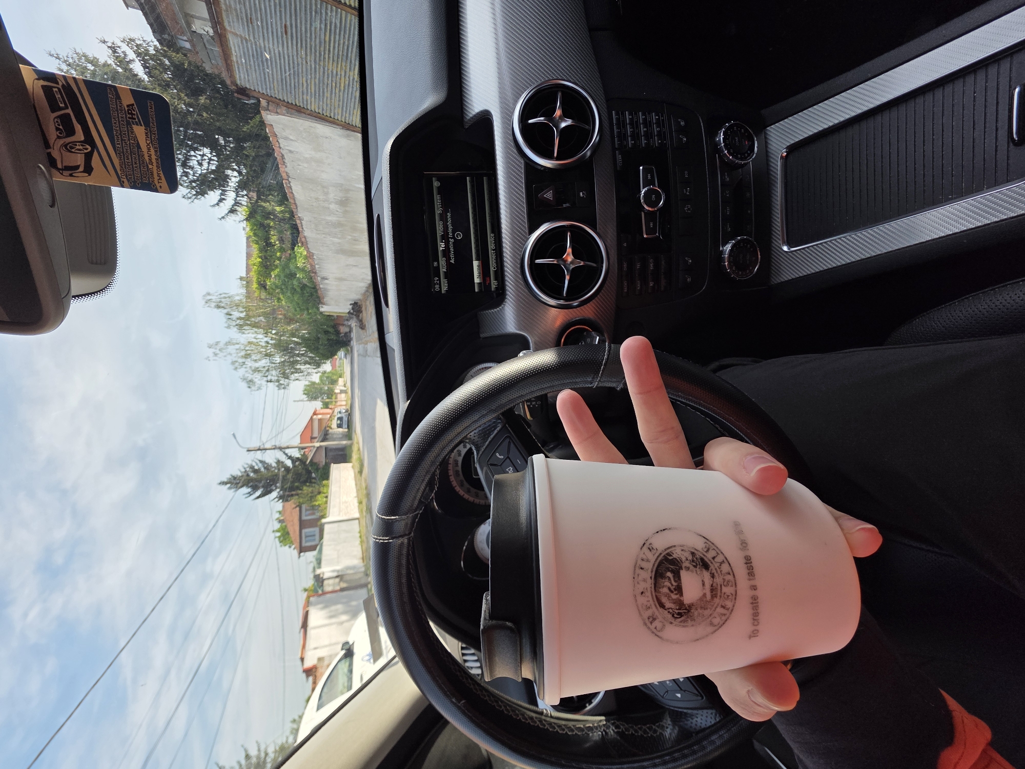 The image captures an interior view from within a car, likely a Mercedes-Benz based on the dashboard features. The steering wheel is in the foreground, behind which lies a view through the windshield. The location seems to be Yagodovo, Bulgaria, based on the vehicle inspection sticker.
A hand holds a white tumbler or mug with a black lid, featuring a logo that reads "CREATIVE CAFE STYLE To create a taste for". The hand is positioned over the steering wheel, with the fingers slightly spread apart.
The car's dashboard is visible with its typical air vents, control buttons, and a display screen showing information like the time (08:29) and various menu options.
Through the windshield, a residential street is visible with houses, trees, and power lines. A car is also visible in the distance.