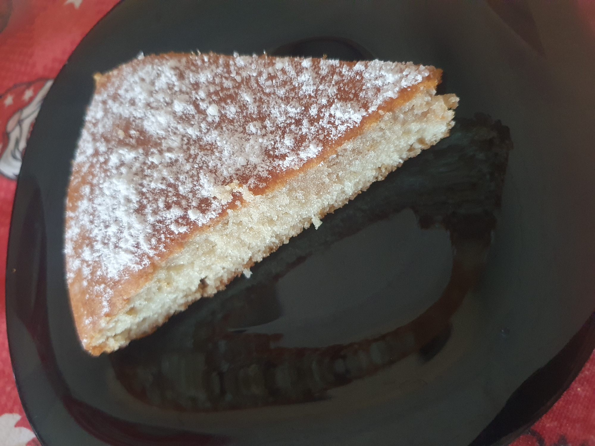 A triangular slice of cake dusted with powdered sugar sits on a glossy black plate. The cake has a slightly browned top and a light-colored, porous interior. The plate reflects an indistinct image, possibly of the surrounding environment. The plate rests on a surface with a patterned red and white covering. The location is unknown.