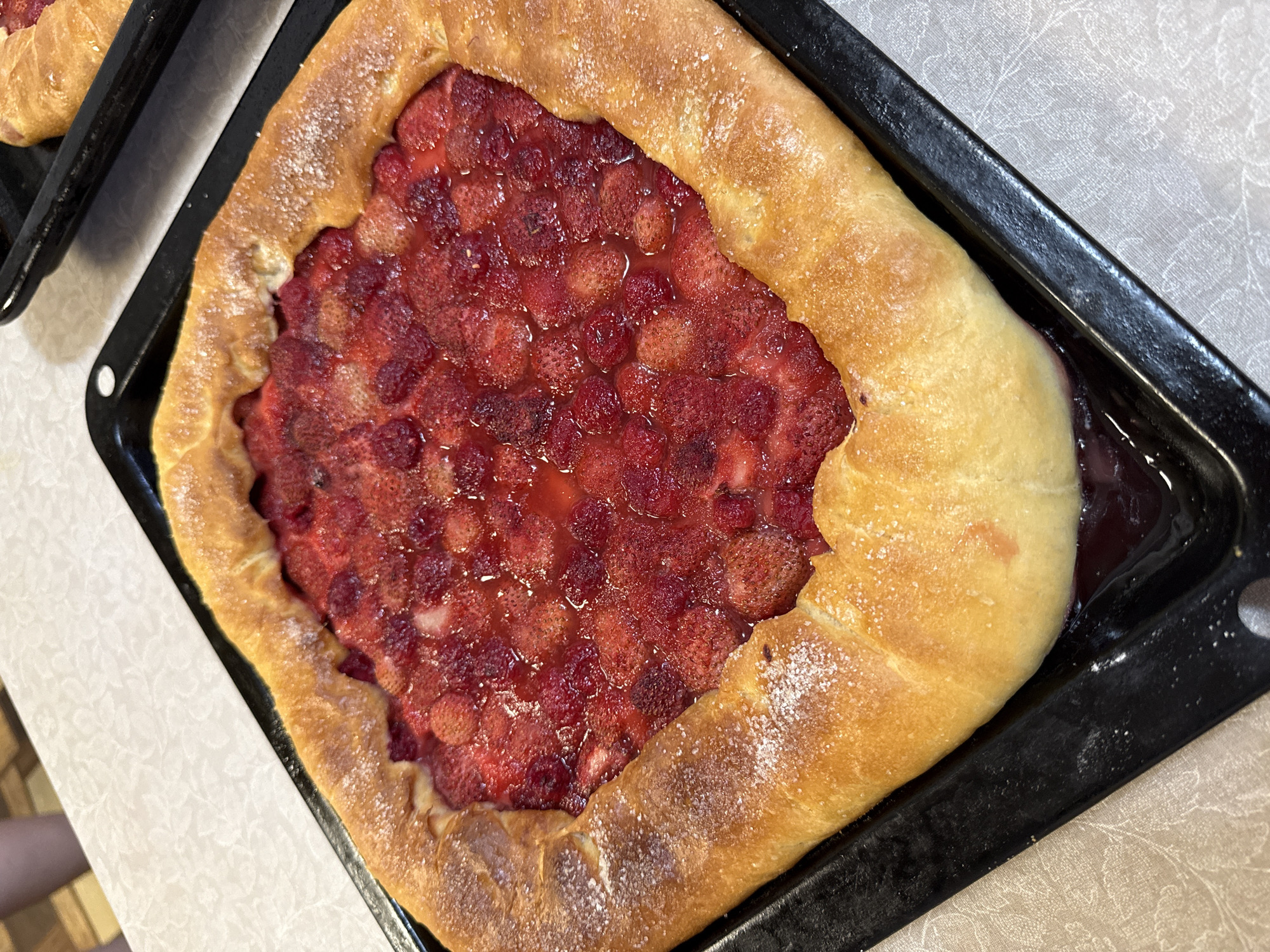 A golden-brown, square-shaped galette filled with halved and whole strawberries rests on a black baking sheet. The pastry is slightly uneven and dusted with sugar, giving it a glistening appearance. The strawberries are a vibrant red, some appearing cooked and slightly softened, with a clear, syrupy liquid surrounding them. The baking sheet is placed on a light-colored, textured surface, likely a kitchen countertop. In the background, the edge of another black baking sheet with a similar pastry is visible, suggesting multiple items were prepared. The overall scene is an indoor domestic setting, possibly a kitchen or dining area, with a portion of a person's legs visible in the upper left corner, indicating human presence. The location is specified as Ветлужский, Russia.
