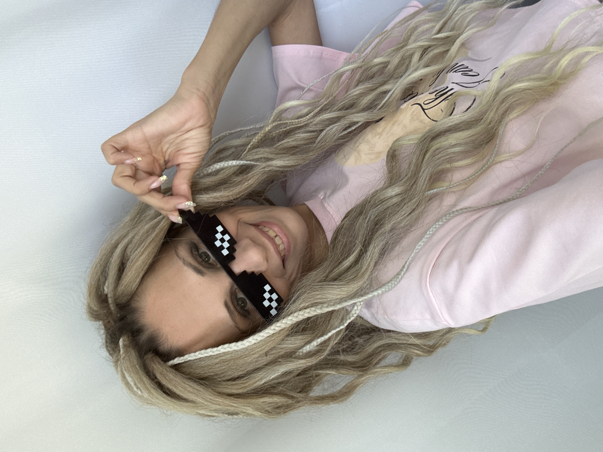 A woman with blonde hair and wearing pixelated sunglasses is looking at the camera. She is wearing a light pink sweatshirt. Her left hand is near her face. The background is a plain white surface. The location is not discernible from the image.