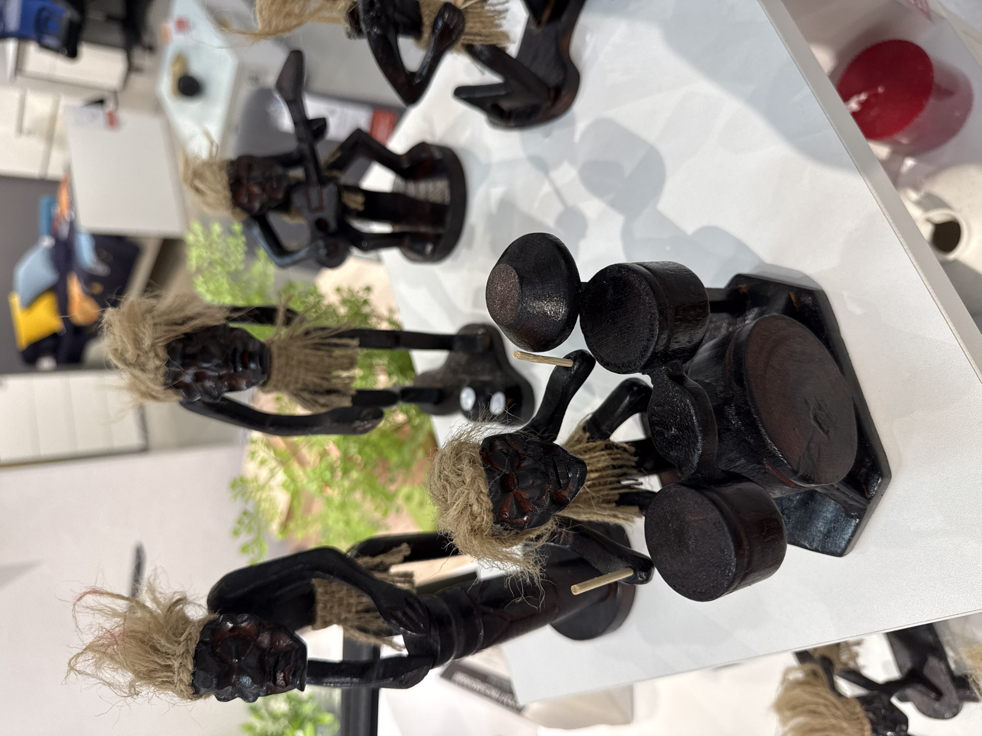 The image shows a collection of carved wooden figurines displayed on a white surface, possibly a shelf or counter. The figurines appear to represent musicians playing various instruments. One figurine plays the drums and another plays the guitar. The figures are dark-stained wood and have accents of light-colored natural fiber representing hair. The setting seems to be a retail environment, possibly a souvenir shop or a store in Perm, Russia. Other items are visible in the background, including household goods like candles and decorative vases. The specific location within Perm is not apparent from the image.
