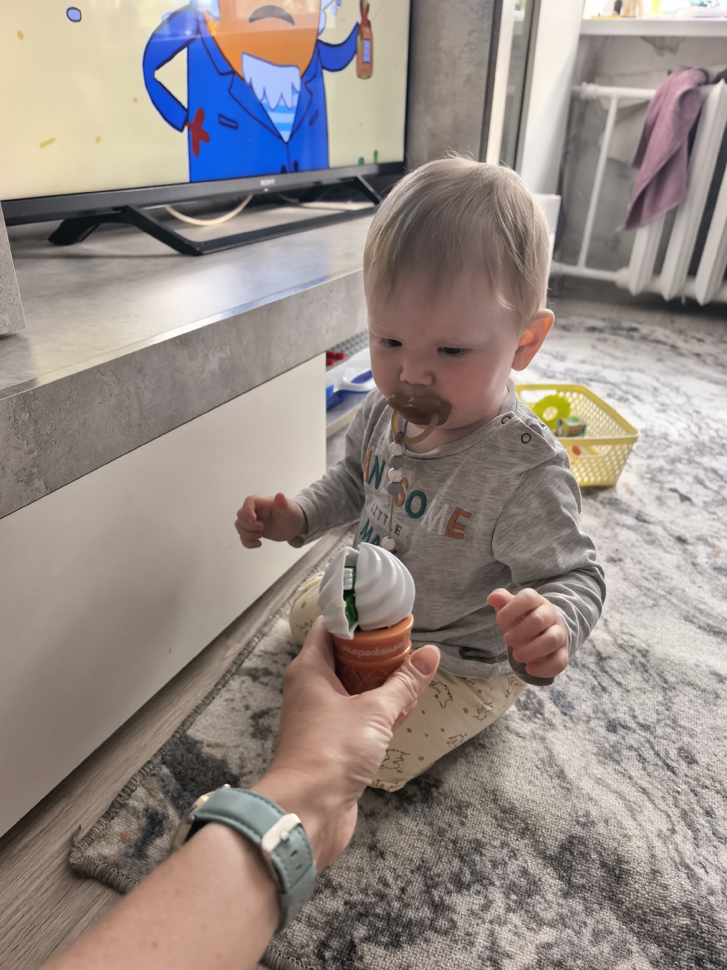 This image depicts an indoor scene in a residence located in Sterlitamak, Russia. A fair-skinned toddler, estimated to be between one and two years old, sits on a grey and white patterned area rug. The toddler has short, light blonde hair and wears a grey long-sleeved shirt with partially visible text "AWESOME LITTLE ME" and light-colored pants with faint animal patterns. A brown pacifier is in the toddler's mouth, attached to a white and grey beaded pacifier clip. The toddler's attention is focused downward on a plastic ice cream cone-shaped toy, white on top and orange on the bottom, being presented by an adult's hand. The adult's left wrist is visible, adorned with a light blue wristwatch featuring a silver buckle.
In the background, a black Sony flat-screen television rests on a grey-topped media console with a white base. The television screen displays an animated character with an orange beard, a blue suit with a red tie or marking, holding a brown bottle against a yellow background. To the far right, a white, ribbed radiator is partially visible, with a purple towel draped over it. Behind the toddler, a yellow plastic basket containing other small toys, including a bright yellow ring, is present on the rug.