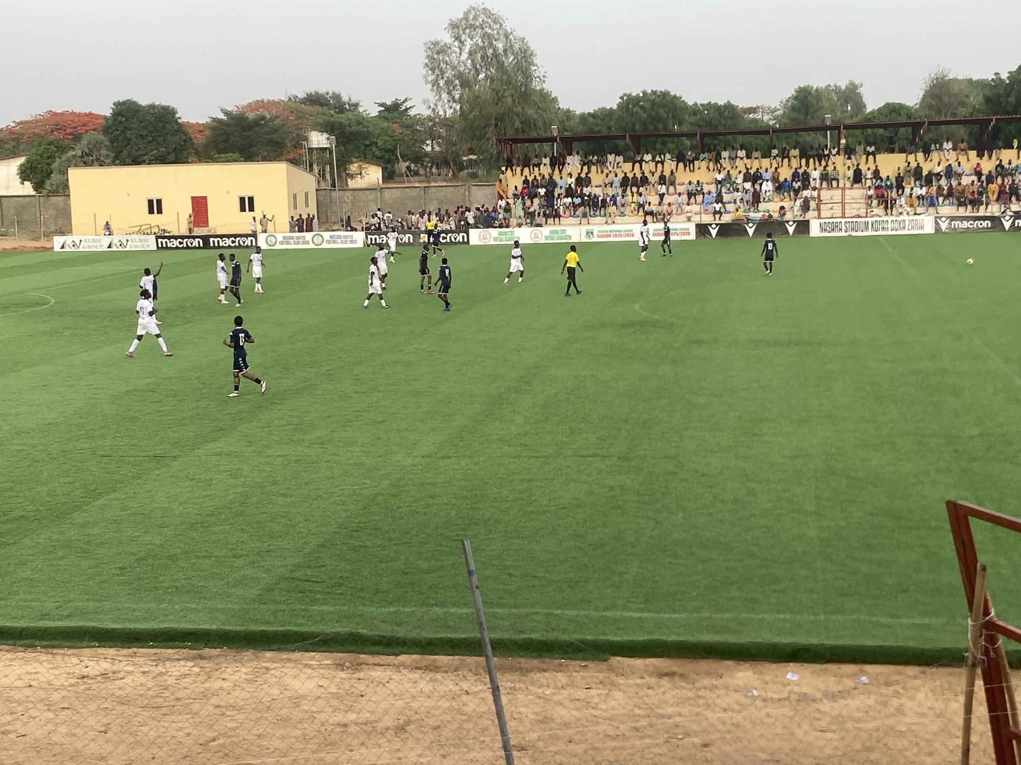 The image shows a soccer game in progress on a green field with spectators in the stands. The banners with Macron are placed along the edge of the field, displaying the text "NASARA STADIUM KOFAR DOKA ZARIA SEASON 2025/2026". The location is Tudun Wada, Nigeria. Players wearing dark blue and white uniforms are running on the field. A small, yellow, rectangular building with a red door is visible near the left side of the frame.