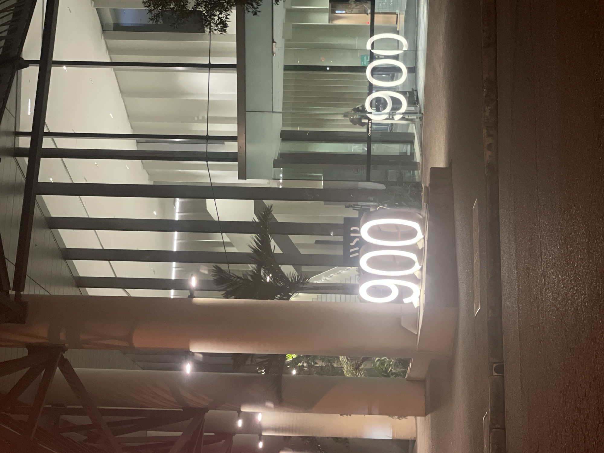 The image depicts the exterior of a modern building at night, with a focus on illuminated numbers displayed prominently. The building features a large glass facade, allowing views into a well-lit interior with vertical structural elements and a high ceiling. In front of the building, large, bright white neon numbers "900" are visible on the left side of the image, and another set of "900" numbers are visible on the right side, displayed on the glass. A palm tree is visible behind the "900" on the left, and another is partially visible on the right. A person is seen through the glass on the right, standing near the entrance. The street in front of the building is paved and appears wet, suggesting recent rain. The architectural style and the presence of tropical foliage, like the palm trees, are consistent with Brisbane, Australia. The lighting suggests it is either early morning or late evening.