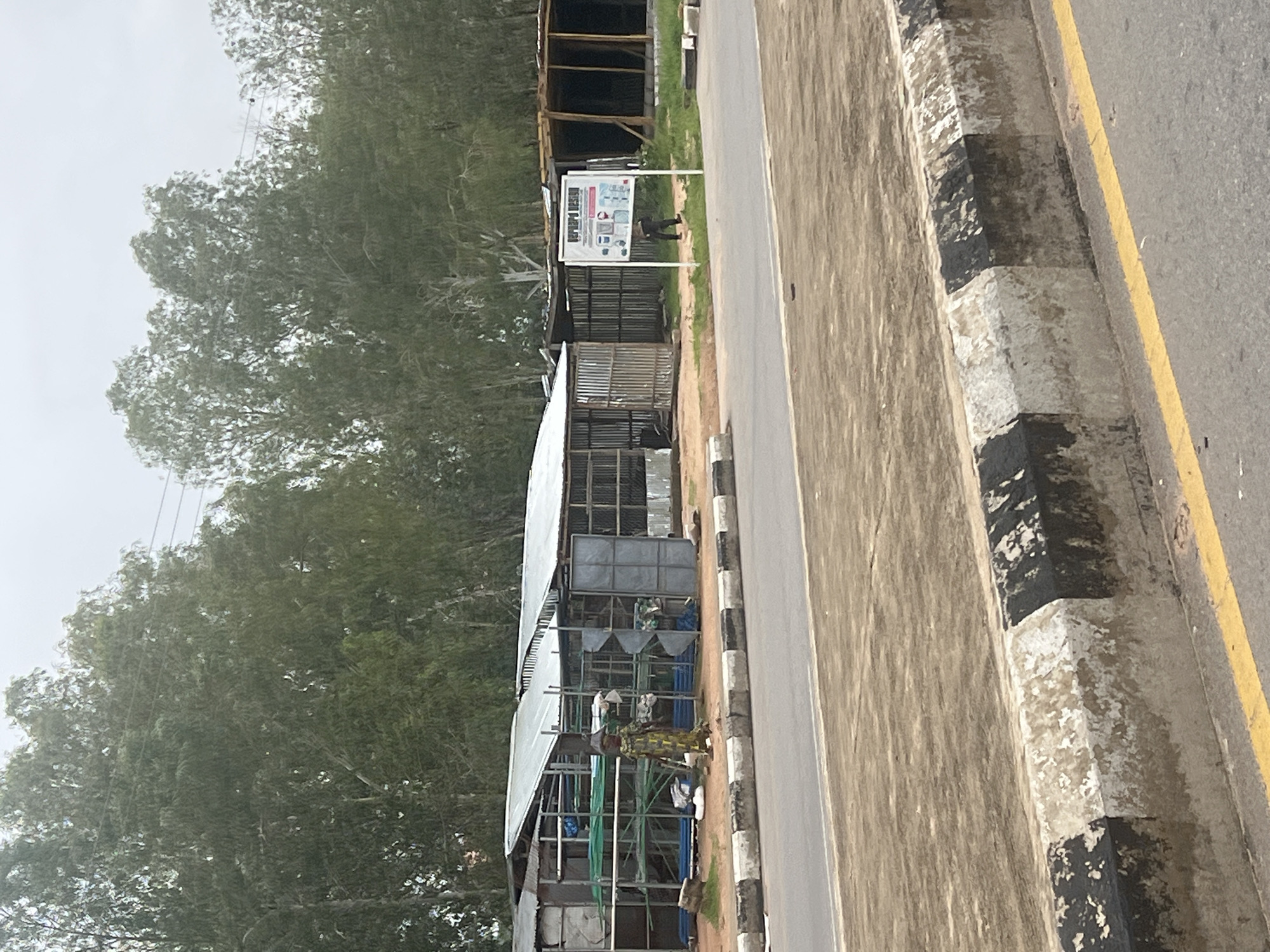 The image depicts an outdoor scene in Jalingo, Nigeria. In the foreground, a road with a yellow line marking the edge of a lane is visible, separated from a dirt embankment by a concrete barrier painted with alternating black and white stripes. Behind the barrier, several open-air stalls are constructed with corrugated metal roofs and sides, some with visible shelving units holding various materials, including what appears to be piping and green sheeting.
A person dressed in a patterned traditional garment stands in front of one of the stalls, facing towards the camera. To the right of the stalls, a white sign with text and images is mounted on a pole. Beyond the stalls, a dense line of tall, green trees forms the background. The sky is overcast, suggesting a cloudy day. A second individual, dressed in dark clothing, is walking away from the sign towards the stalls. The overall impression is a roadside marketplace or storage area.
