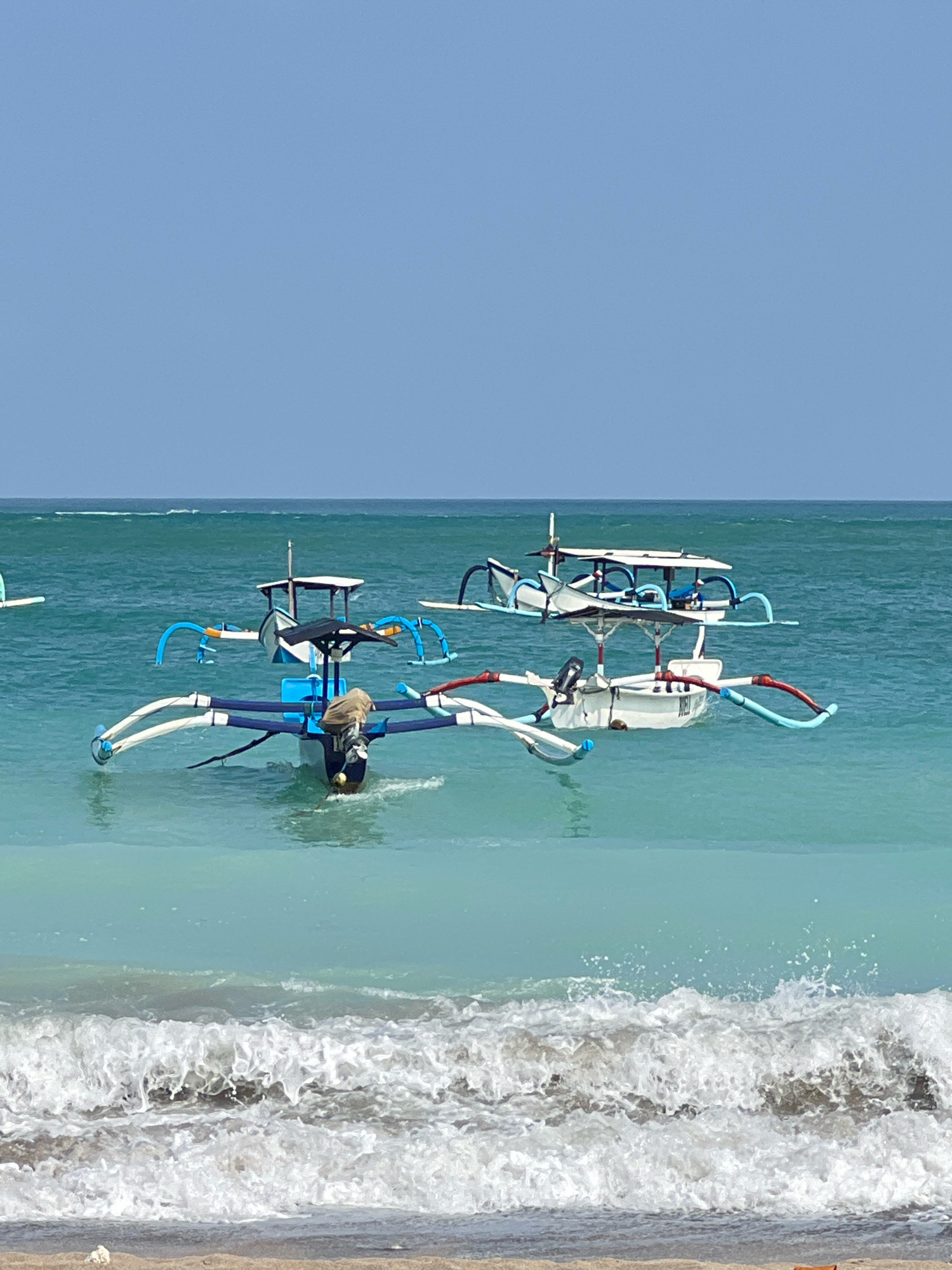 In Kuta, Indonesia, two outrigger boats are moored in the turquoise waters of the ocean. The boat in the foreground is blue and white, with a brown tarp draped over its engine. The second boat, further out, is white with blue accents. Waves are breaking on the sandy shore in the foreground, with the blue sky visible above the horizon.