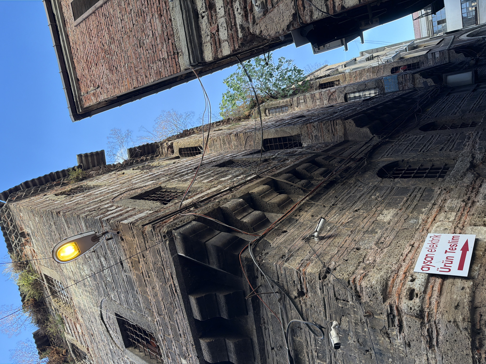 The image shows a street in the Beyoğlu district of Istanbul, Turkey. The scene is dominated by two old, brick buildings facing each other at an angle, creating a narrow alleyway. The building on the left is constructed from dark stone and brick, with visible wear and tear, and several windows with metal bars. A streetlamp with a bright yellow light is mounted on this building. Wires are strung across the alleyway. A white sign with red text and an arrow pointing to the right is attached to the lower part of this building. The sign reads "Aysan Elektrik Ürün Teslim". On the same building, there are at least two white security cameras mounted. The building on the right is made of red brick and appears to be in slightly better condition, also with barred windows. Vegetation, including trees and bushes, is growing from the tops and in between the buildings. The sky above is clear blue. No people are visible in the image.