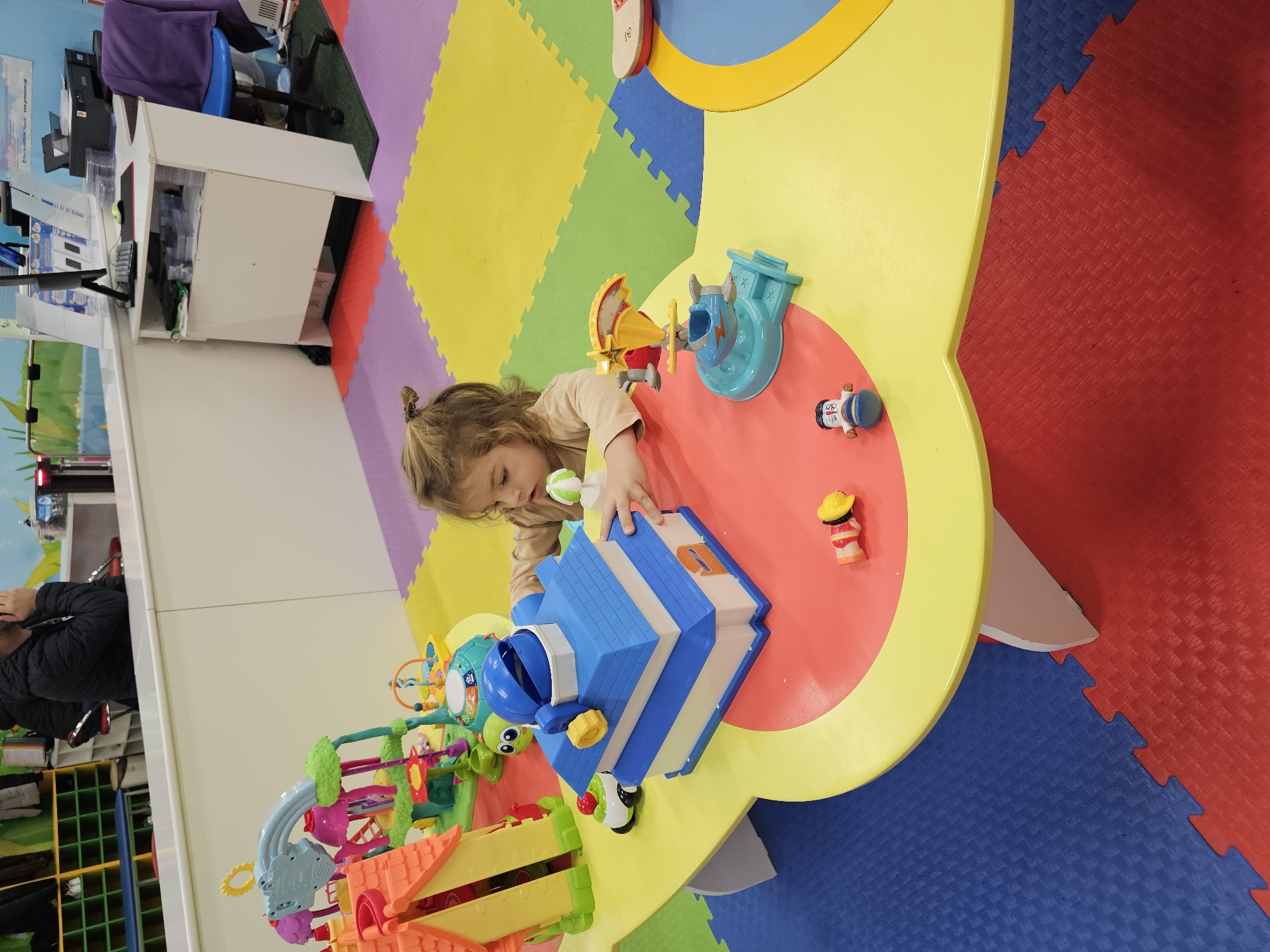 A child with blonde curly hair is engrossed in playing with toys on a multi-colored play table. The table has a bright yellow, flower-like shape with a red inner circle. On the table, there are various toy sets, including a blue house-shaped toy, a yellow and red windmill-like toy, and several small figurines. The child is wearing a light brown shirt and seems to be focusing intently on the toys.
The surroundings feature colorful interlocking floor mats in yellow, green, purple, red, and blue, providing a safe play area. In the background, there's a white cabinet, and the location appears to be an indoor play area, possibly a children's center, in Khimki, Russia.