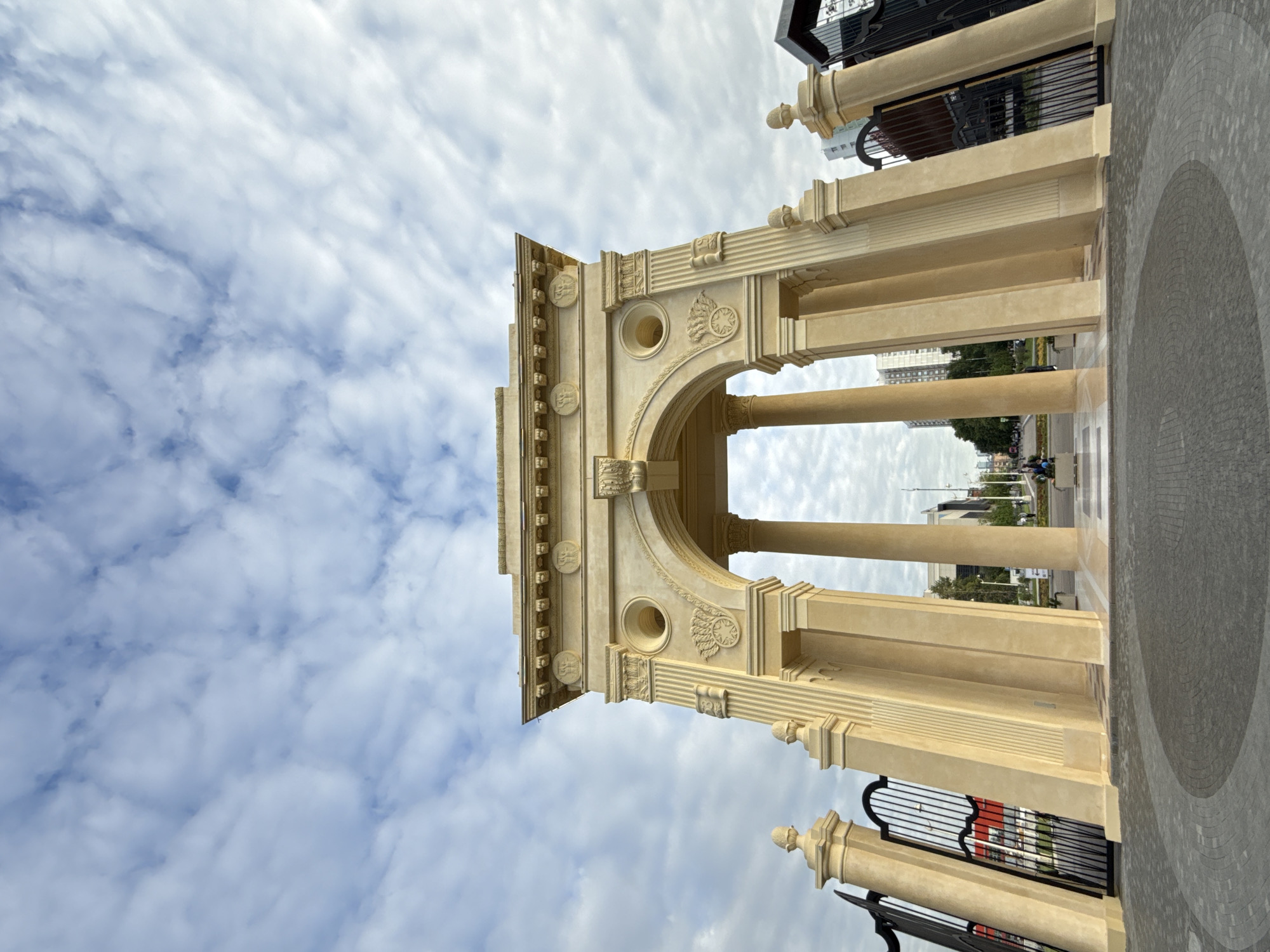 The image captures a grand, ornate archway or gateway, primarily composed of light beige or sand-colored stone. The architecture features classical elements, including fluted columns, detailed cornices, and decorative carvings such as circular medallions with abstract symbols and a prominent round opening within the arch's structure. Integrated into the sides of the gateway are black metal fences or gates with vertical bars and scrollwork.
The archway stands against a sky filled with layered, textured clouds, possibly altocumulus, indicating a partly cloudy or overcast daytime. Beyond the arch, a paved ground with a semi-circular pattern leads towards what appears to be a street or pathway. In the distance, trees line the street, and several modern, multi-story buildings rise against the horizon. Small, indistinct figures of people can be seen in the far background, suggesting a public area. The lighting suggests it is daytime, possibly late morning or early afternoon, with diffused light due to the cloud cover. The style of the archway, combined with the context of Moscow, Russia, suggests a neo-classical or monumental public structure. No legible text is visible in the image.