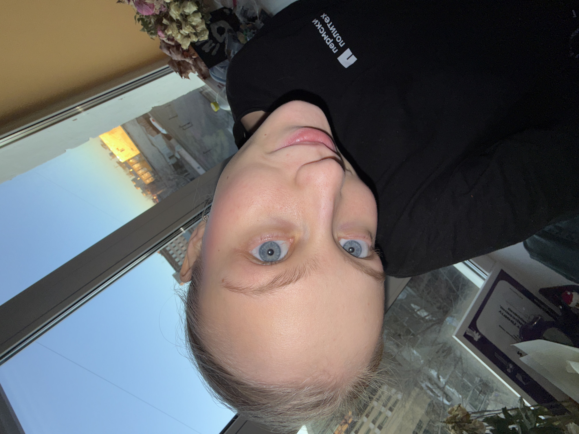 A selfie-style photograph shows a woman indoors, oriented with her head at the bottom of the frame. She has fair skin, blue eyes, and light blonde hair. She is wearing a black top with Cyrillic text and a logo that reads "Пермский политех." The background features a window with a reflection of a city scene at sunset, possibly Perm, Russia, based on the text on her top and the urban architectural style. Near the window, a framed document and some dried flowers can be seen.