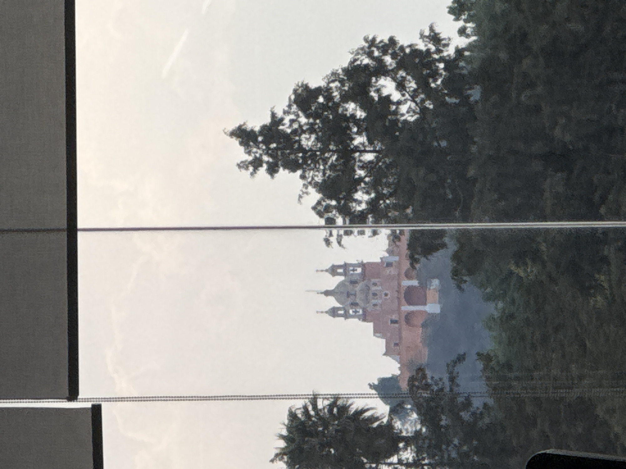 The image shows a view of the Santuario de la Virgen de los Remedios, a church located atop the Tláchicualtepetl pyramid in San Andrés Cholula, Mexico, as seen through a window with blinds. The building appears to be constructed primarily of red-orange stone or brick, featuring arches, domes, and multiple levels. It is partially obscured by trees. The sky is overcast. Window frame and blinds are visible in the image's periphery.