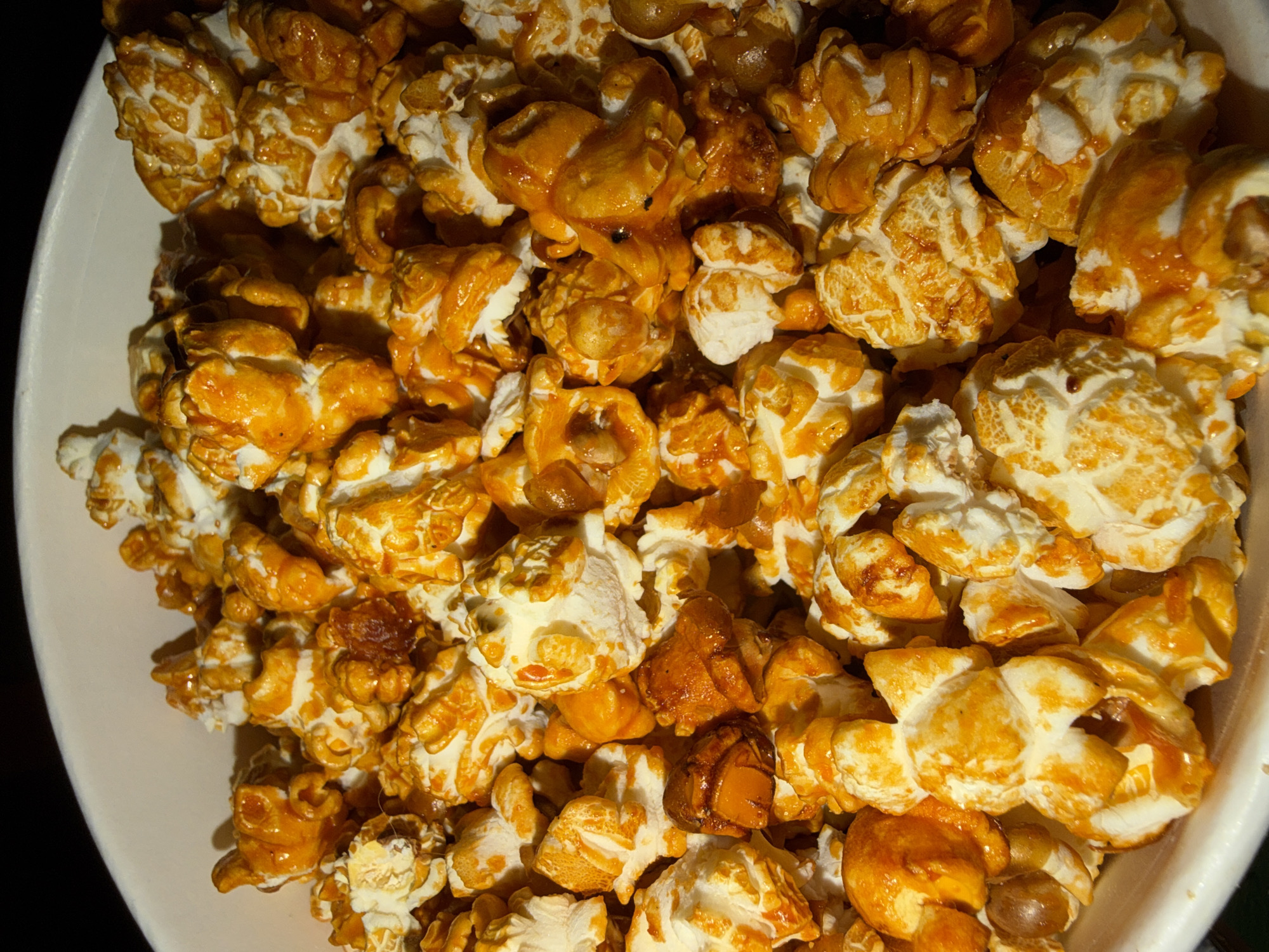The image depicts a close-up of caramel popcorn in a white bowl. The popcorn is a mix of light and dark caramel coating. The scene appears to be indoors with dark surroundings. There's no indication of Chaikovsky, Russia.