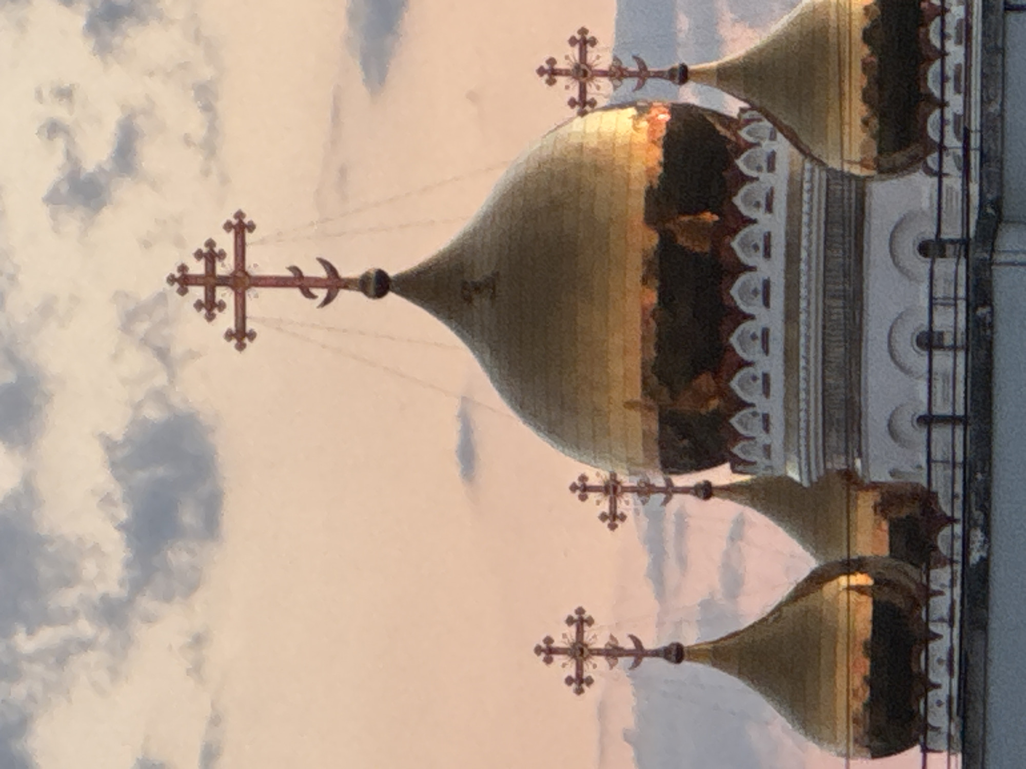 A close-up, vertical, eye-level shot captures the golden domes and crosses of a church against a pale pink and grey sky. The sky is filled with soft, diffused clouds. The main focus is the central, largest dome, which is a bright gold and reflects the sky's colors. It is topped with a cross that has a crescent moon at its base, a common symbol in Eastern Orthodox Christianity. Smaller, similarly designed domes and crosses are visible on either side of the central dome. The domes are ornate, with decorative white trim along their bases. The building appears to be made of stone and metal. The image is taken from a slightly elevated perspective, looking up towards the architectural features.